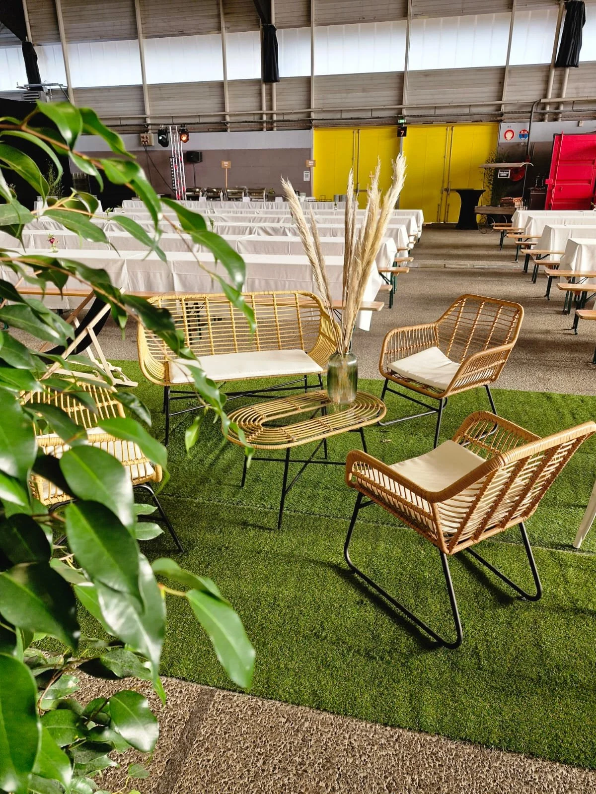 Espace de décoration avec un tapis vert, fauteuils en rotin, une table avec un vase de plumes, une salle avec tables et chaises blanches.