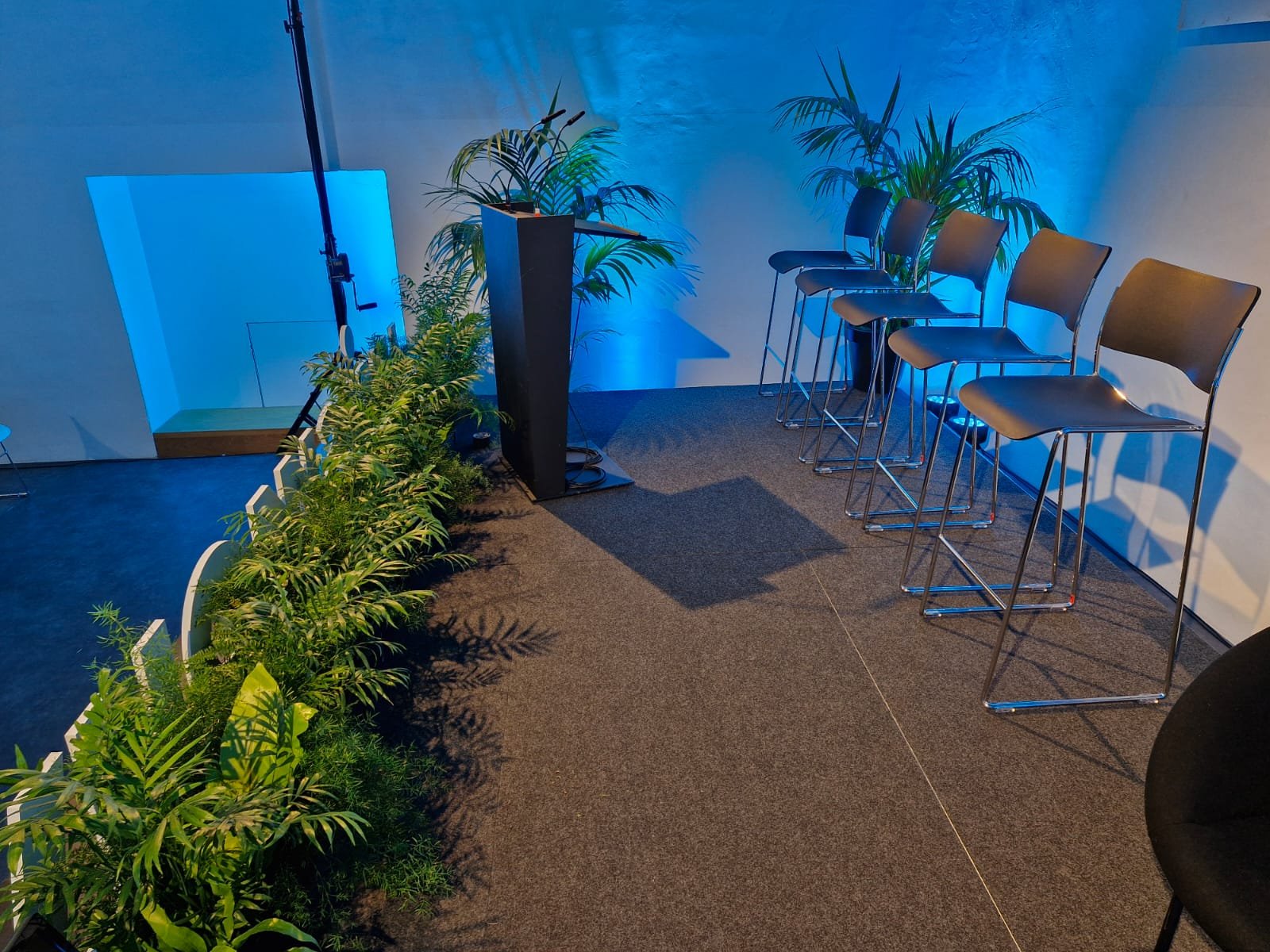 Une scène intérieure avec des chaises en rangée, des plantes vertes, un podium, et un éclairage bleu, suggérant une salle de conférence ou un événement.
