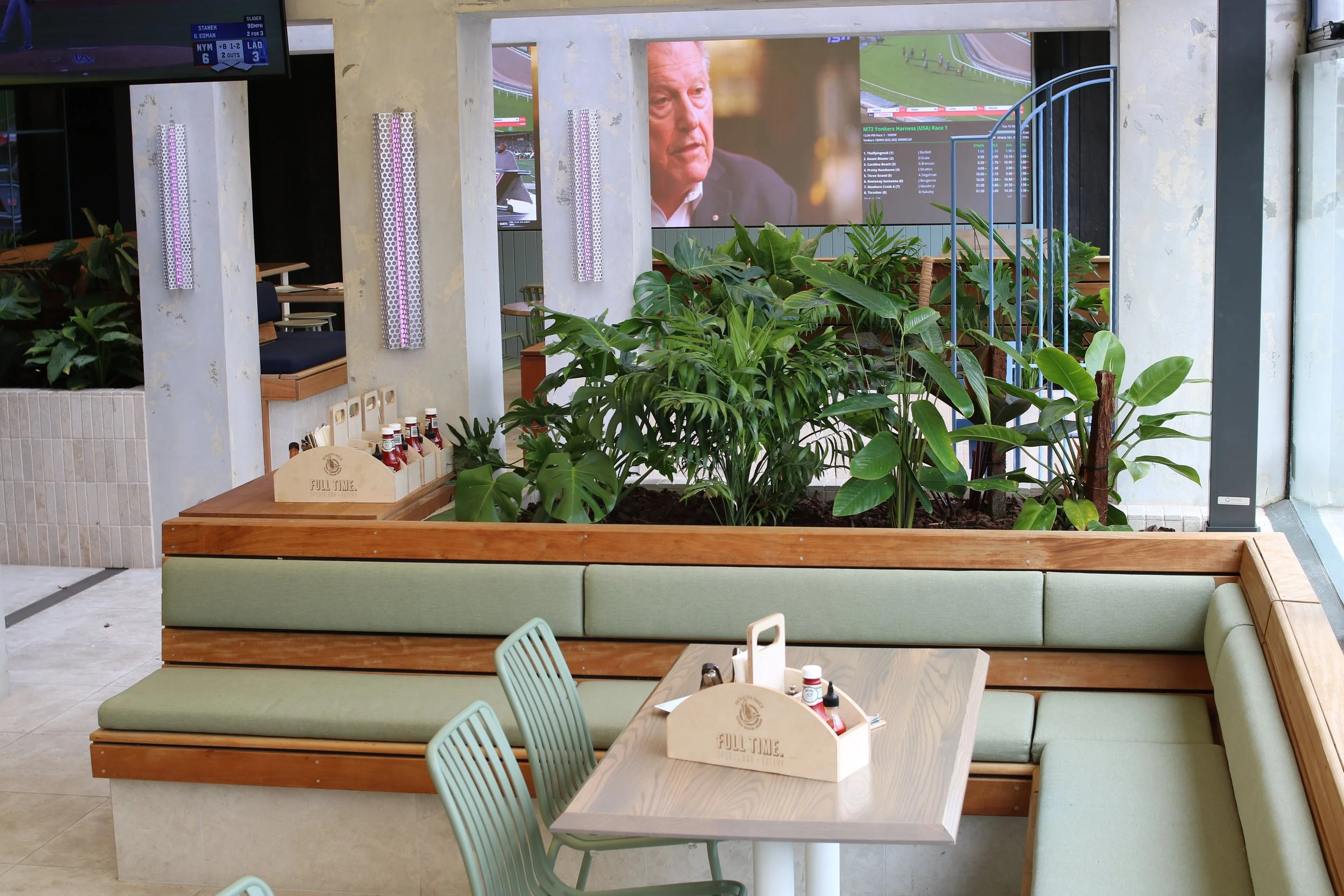Intérieur d'un restaurant ou café avec des tables en bois, chaises en métal, un espace vert avec des plantes, et des écrans de télévision avec un programme de courses hippiques.