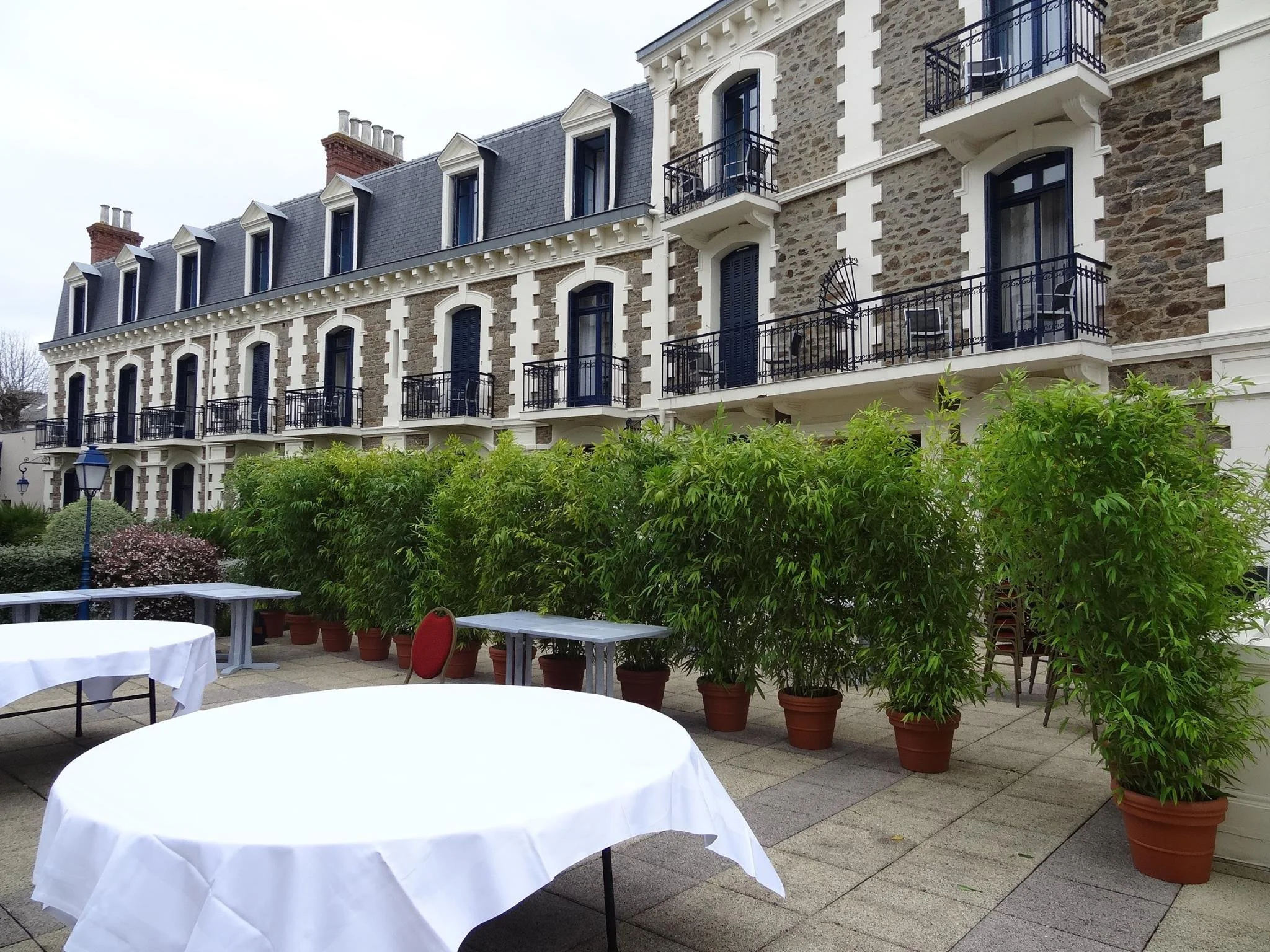 Une terrasse extérieure avec des tables rondes couvertes de nappes blanches. Il y a plusieurs pots avec des arbustes verts. En arrière-plan, un immeuble en pierre avec plusieurs balcons en fer forgé.
