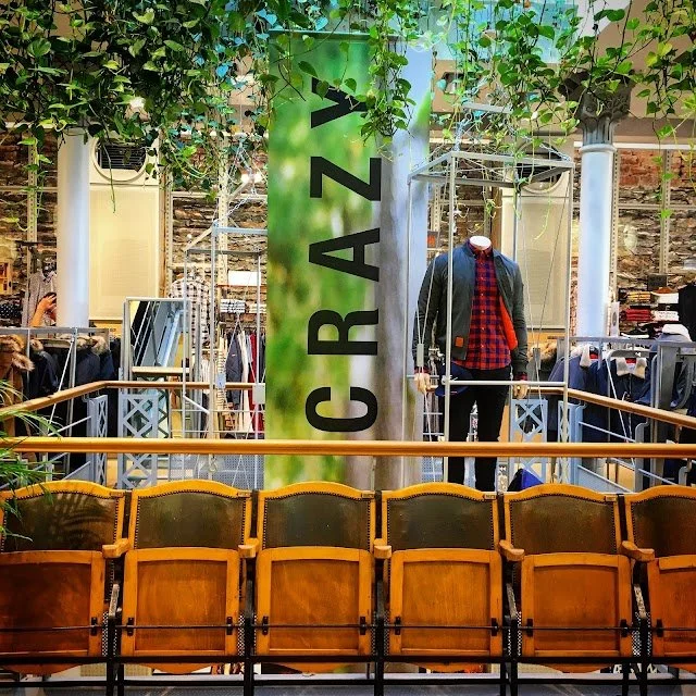 Intérieur d'un magasin H&M avec une rangée de chaises en bois à l'avant, un mannequin portant une veste et une chemise à carreaux, et une bannière verte avec le texte 'Crazy' suspendue au plafond.