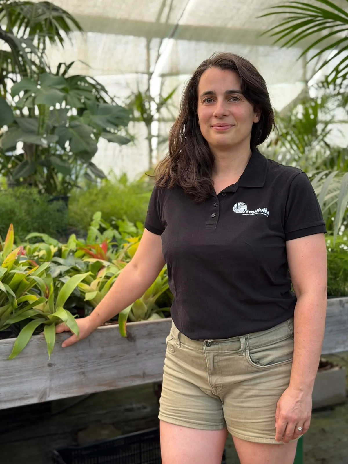 Une femme avec des cheveux bruns, portant un polo noir avec un logo, dans une serre entourée de plantes vertes.