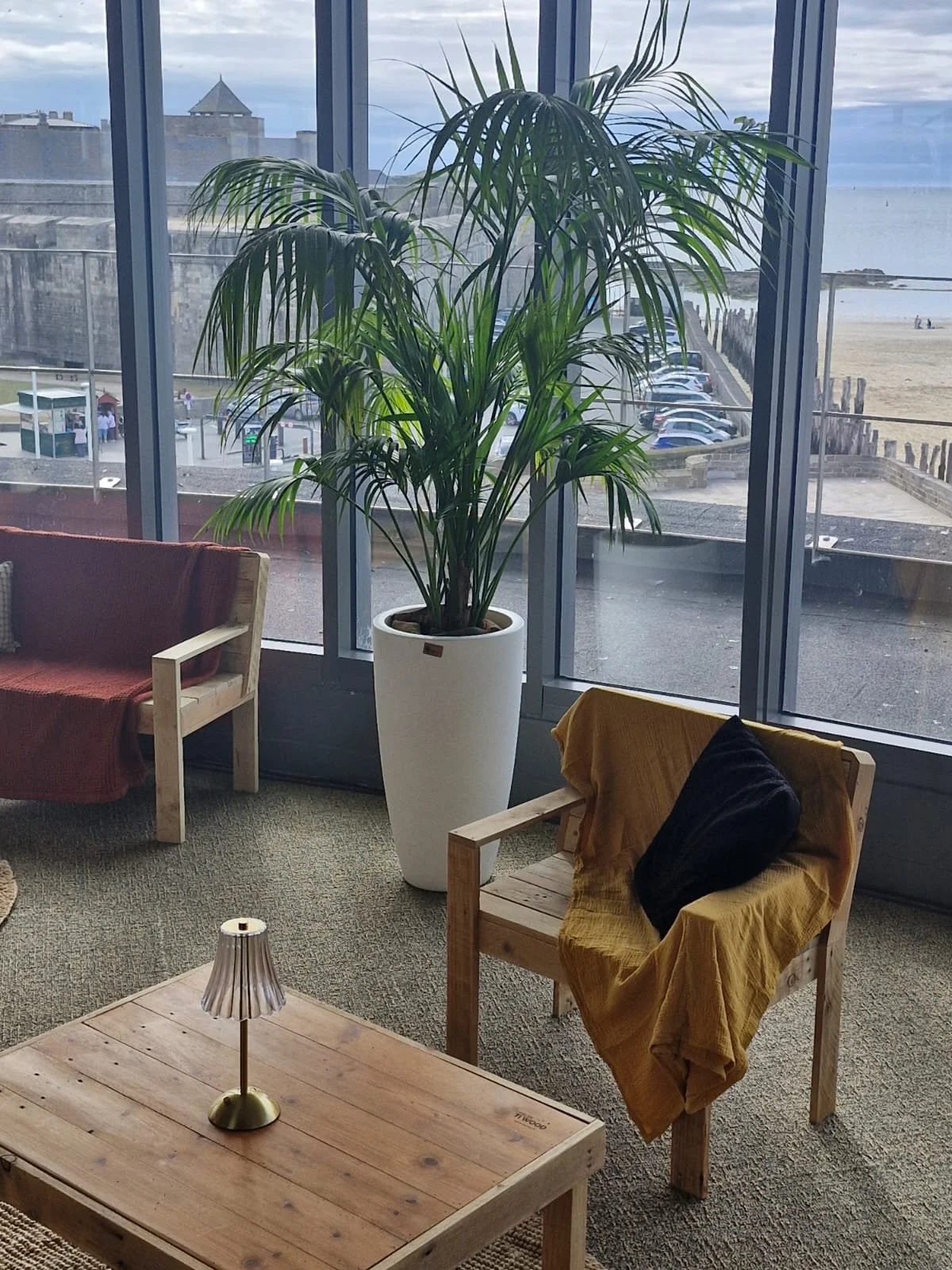 Salon avec une grande plante verte dans un pot blanc, deux fauteuils en bois avec des coussins, une table en bois avec une petite lampe, devant une grande fenêtre qui offre une vue sur la plage et la mer.