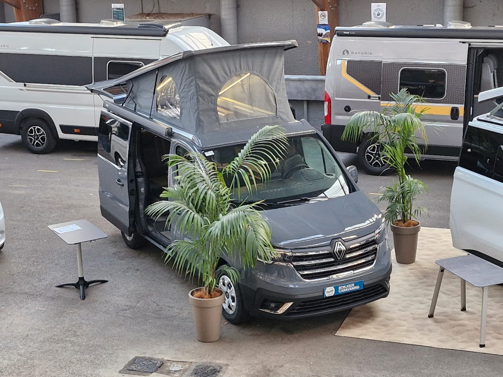 Un camping-car gris avec un toit relevable et des plantes en pot dans un espace intérieur d'exposition. Autres camping-cars en arrière-plan.