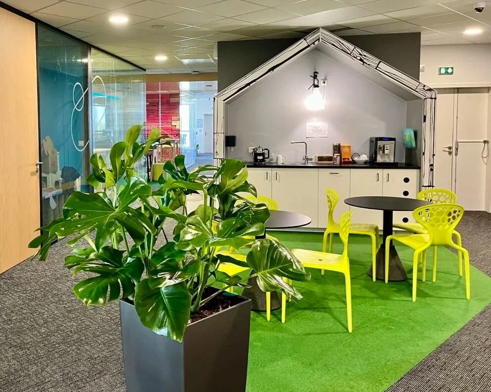 Espace de détente avec une grande plante en pot vert et un coin cuisine avec un petit salon, chaises jaunes, tables noires, et un décor intérieur moderne.