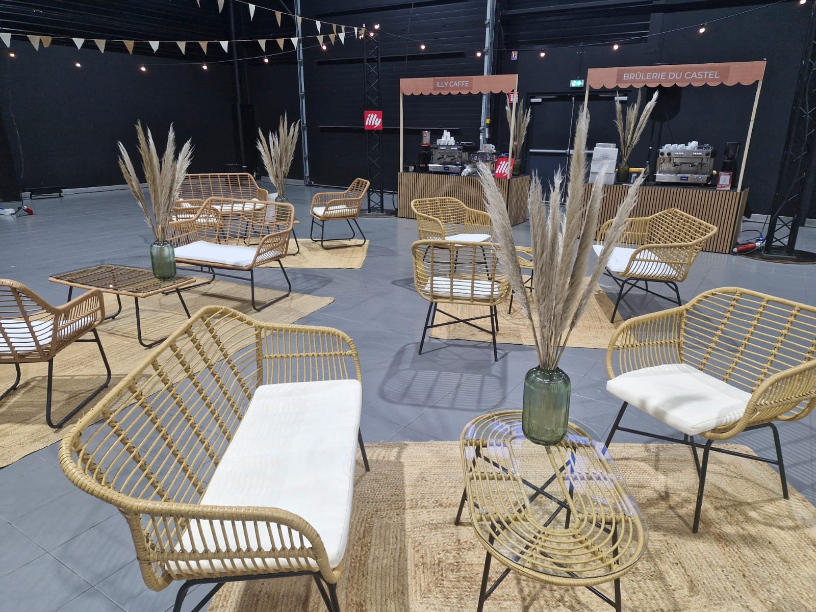 Espace de dégustation avec plusieurs fauteuils en rotin, tables basses en métal, vases avec des pampas et stands de café en arrière-plan dans un décor moderne et épuré.