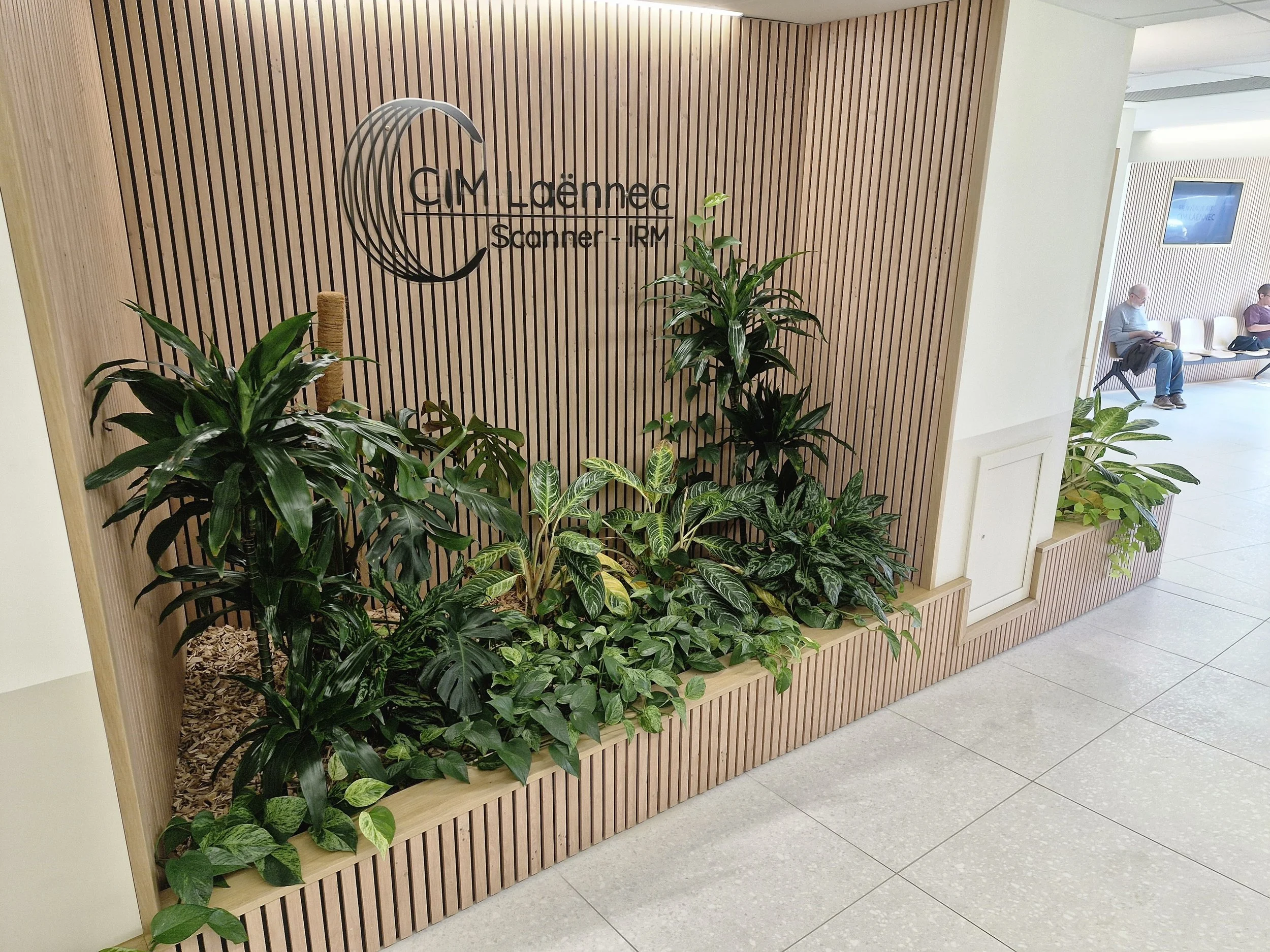 Un mur en bois avec un logo et du texte indiquant 'CIM La Médec, Scanner - IRM'. En dessous, des plantes vertes décoratives sont placées, et à droite, deux personnes sont assises sur une rangée de chaises dans une salle d'attente.