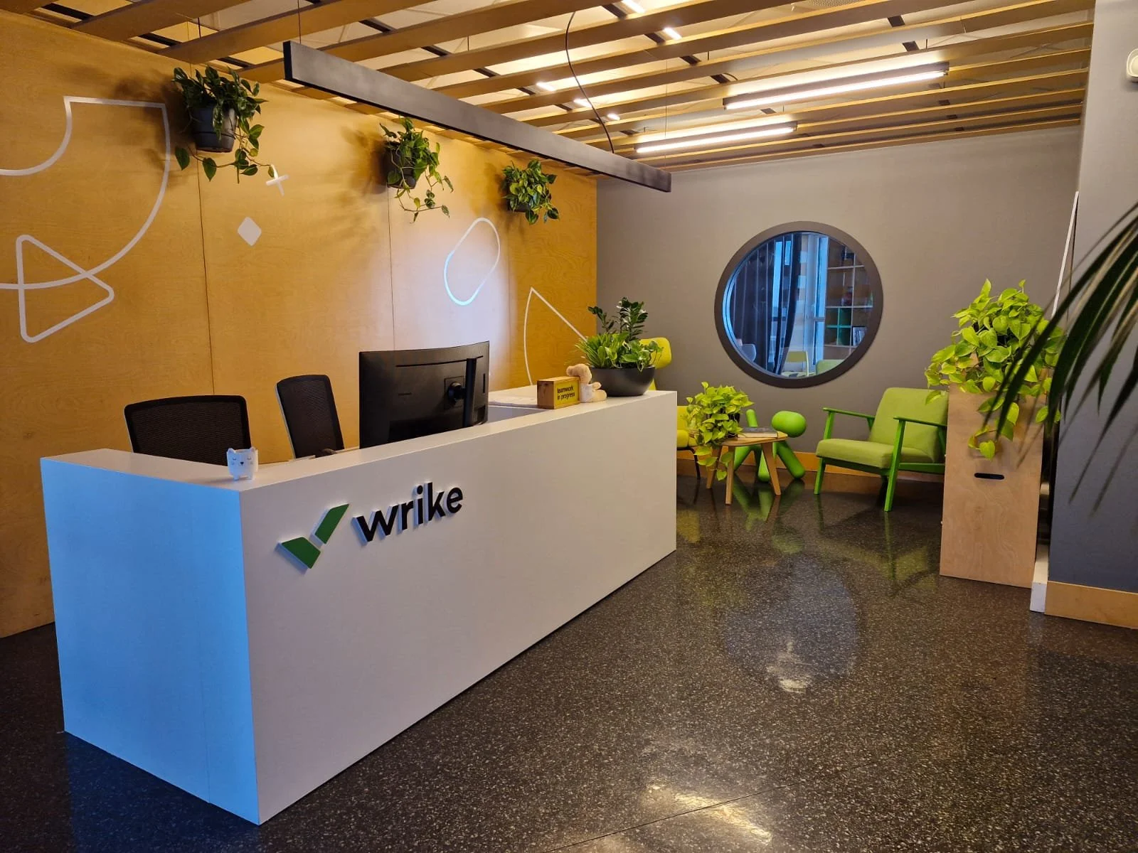Un espace de réception moderne avec un bureau blanc portant le logo 'wrike', des chaises vertes et un mur en bois avec des plantes suspendues. Une fenêtre ronde offre une vue sur un coin salon avec des fauteuils verts.