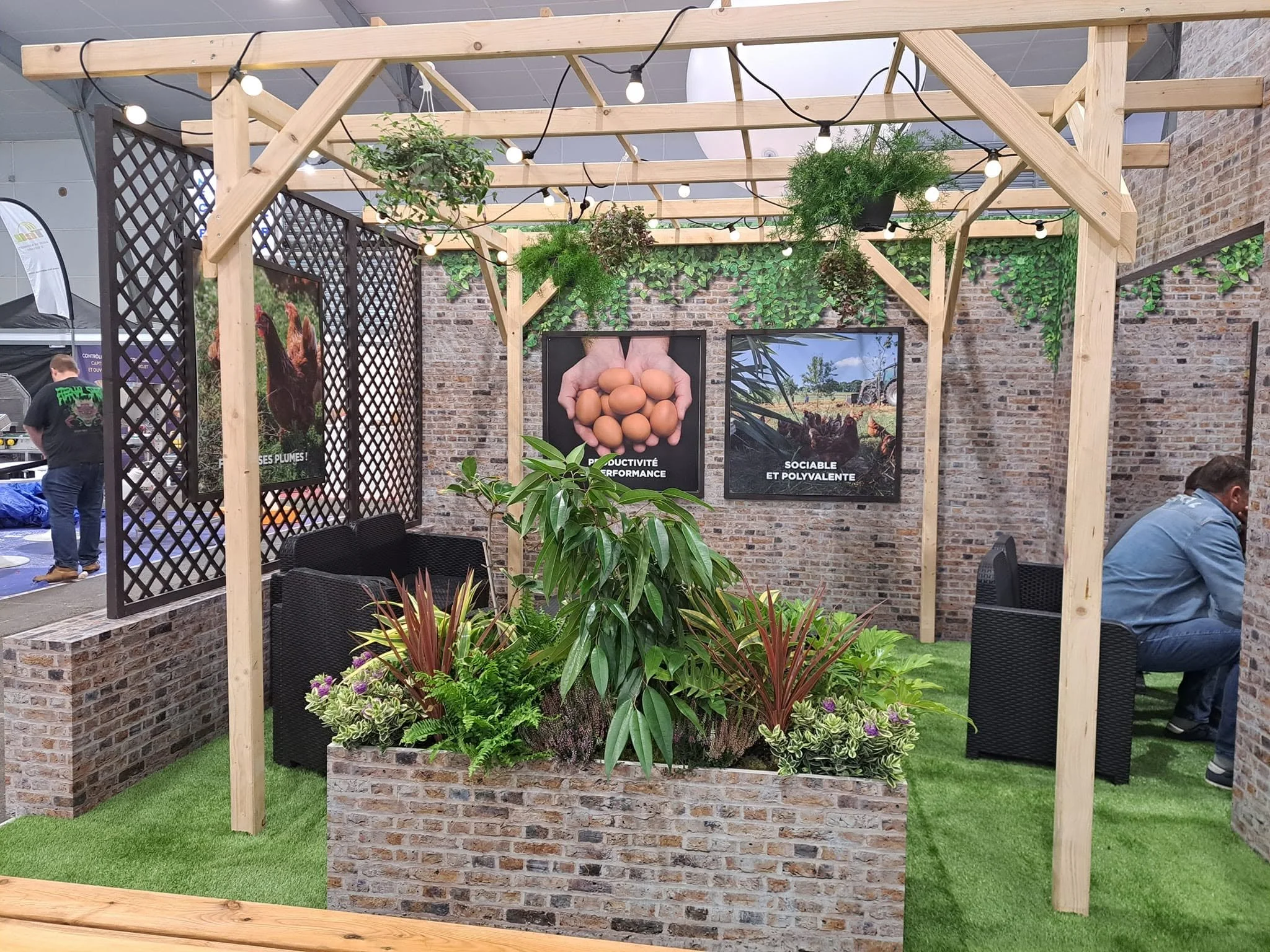 Structure en bois avec des plantes vertes suspendues et panneaux d'affichage, décorée de guirlandes lumineuses, dans un espace extérieur avec des murs en briques.
