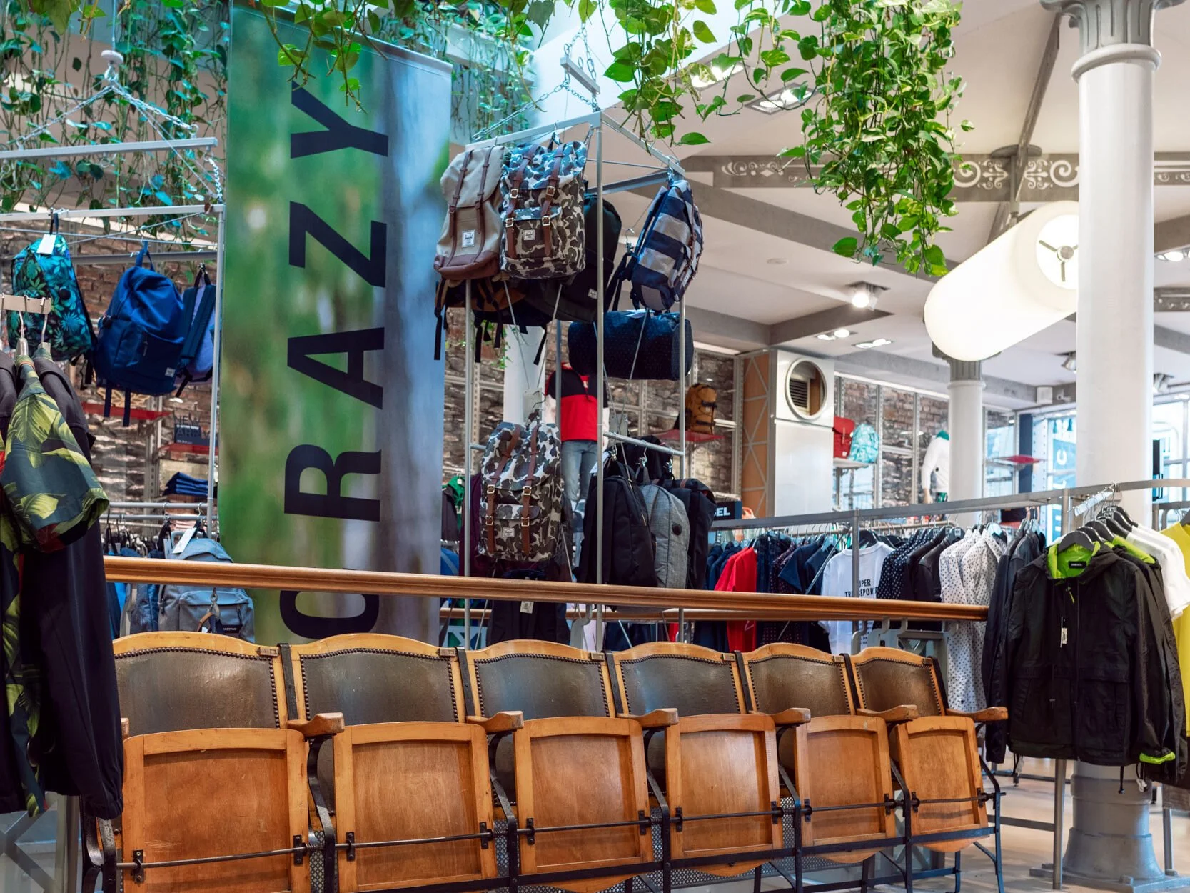 Rayonnage avec sacs à dos et vêtements dans un magasin de sport, intérieur lumineux avec plantes suspendues au plafond.