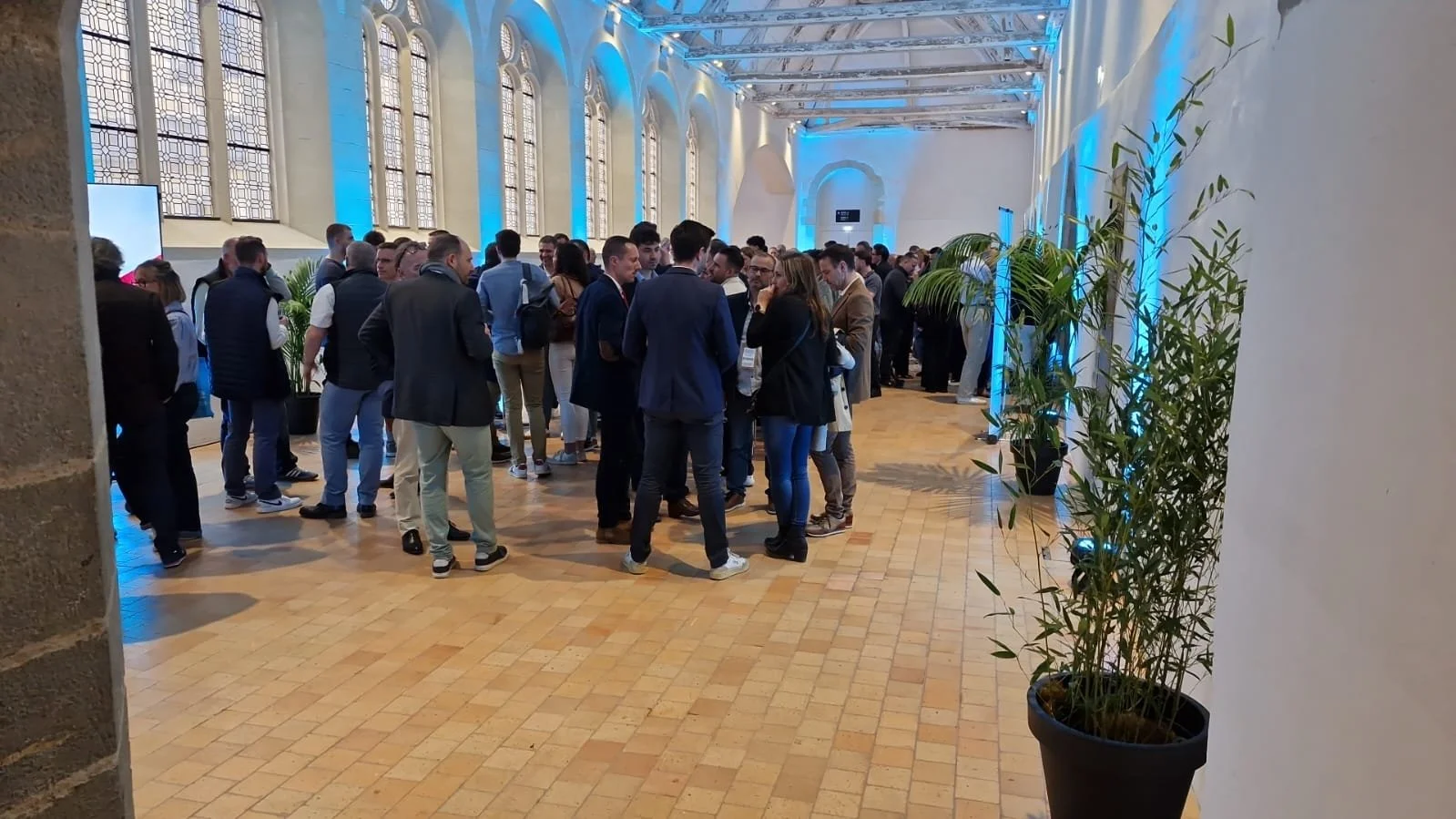 Evénement professionnel dans une salle avec de grandes fenêtres et une lumière bleue. Plusieurs personnes discutent en petits groupes.