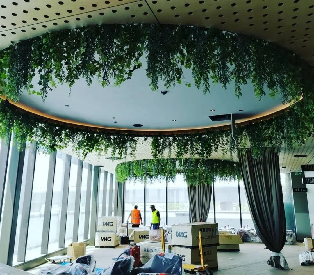 Construction intérieure avec plafond suspendu vert et plantes suspendues, deux ouvriers en gilets de sécurité, grandes fenêtres avec vue extérieure, cartons et matériaux de construction.