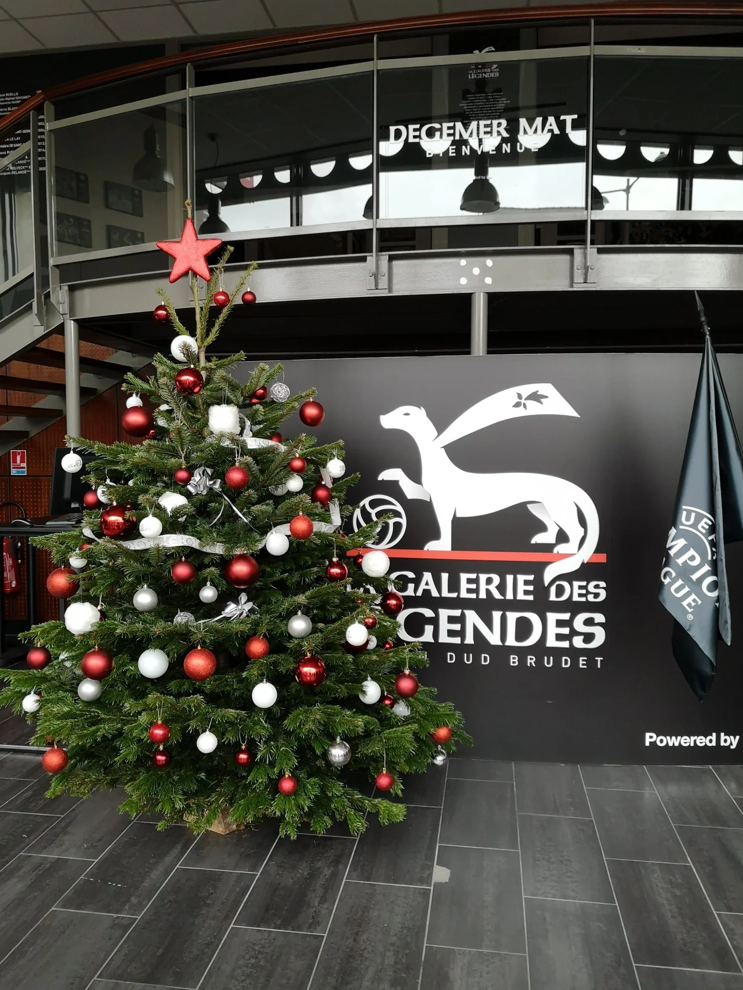 Un sapin de Noël décoré avec des boules rouges, blanches et argentées, et une étoile rouge en haut. En arrière-plan, une façade avec des inscriptions. Le sol est en carreaux gris foncé.