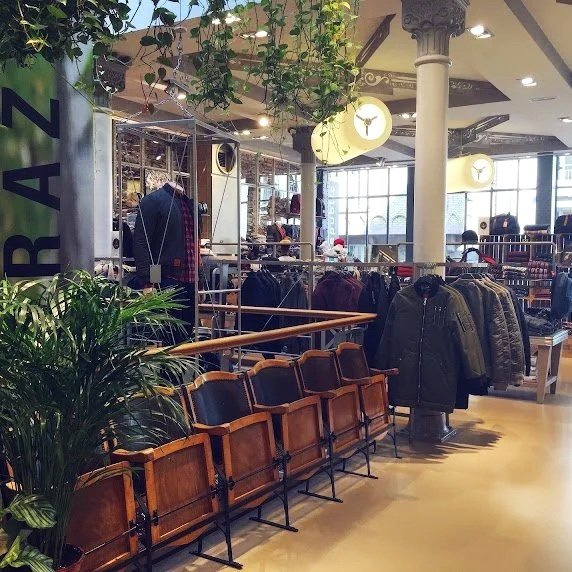 Intérieur d'un magasin de vêtements avec des chariots, des hommes portant des manteaux, des étagères de vêtements, des plantes et des horloges au plafond.