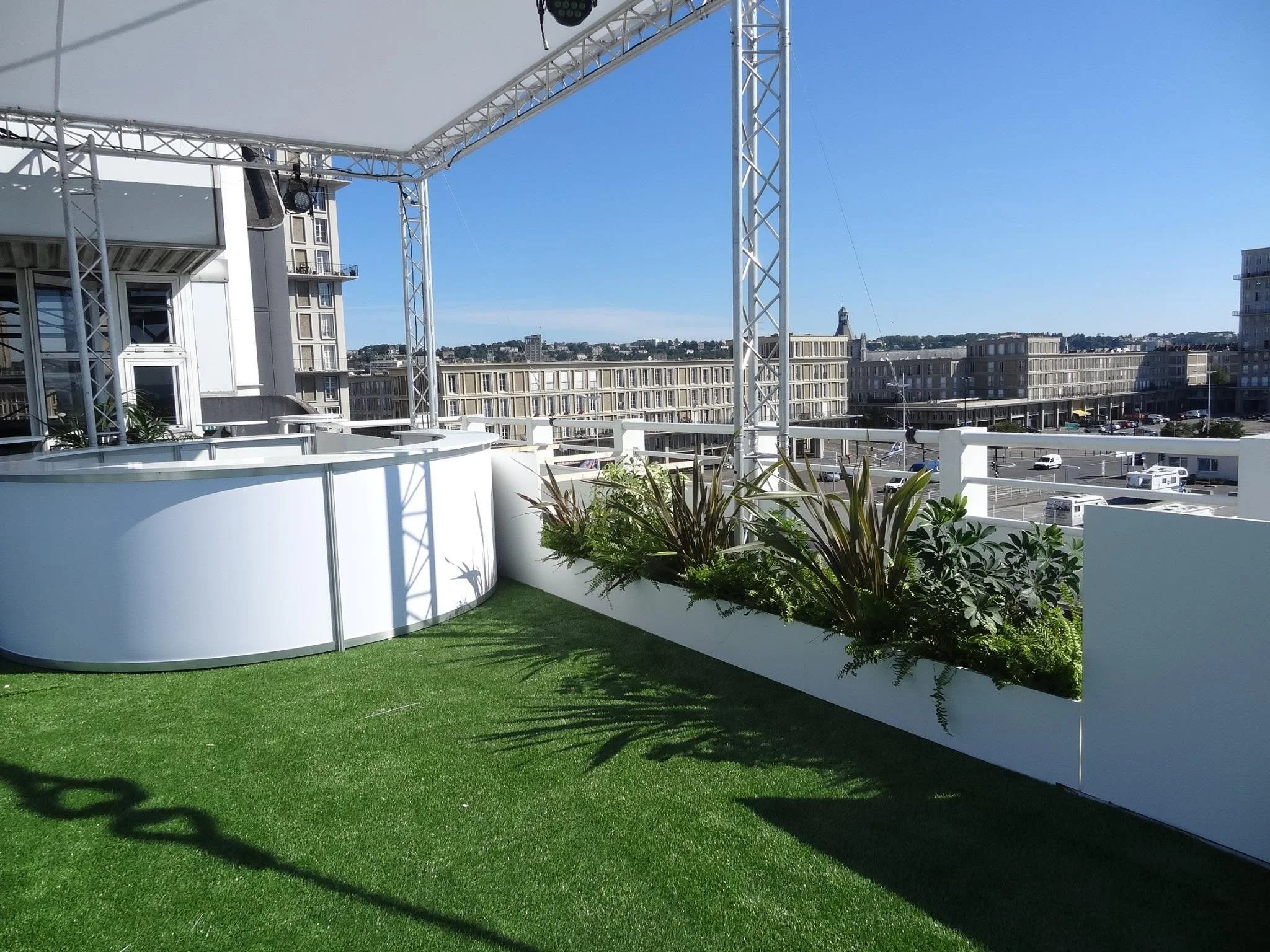 Une terrasse avec un sol en herbe artificielle, entourée de barrières blanches, avec des plantes en pot. En arrière-plan, des bâtiments modernes et un ciel dégagé.