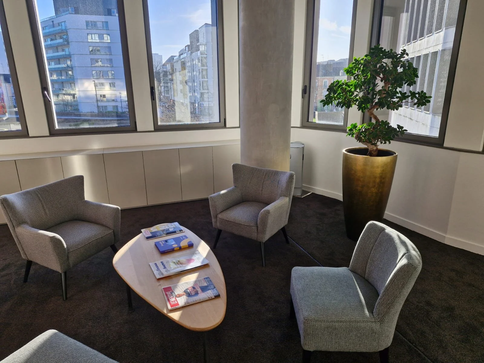 Salle de réunion ou salon avec quatre fauteuils gris autour d'une table en bois, magazines dessus, grand pot de fleurs avec un arbre, fenêtres avec vue sur des bâtiments modernes, intérieur bien éclairé.