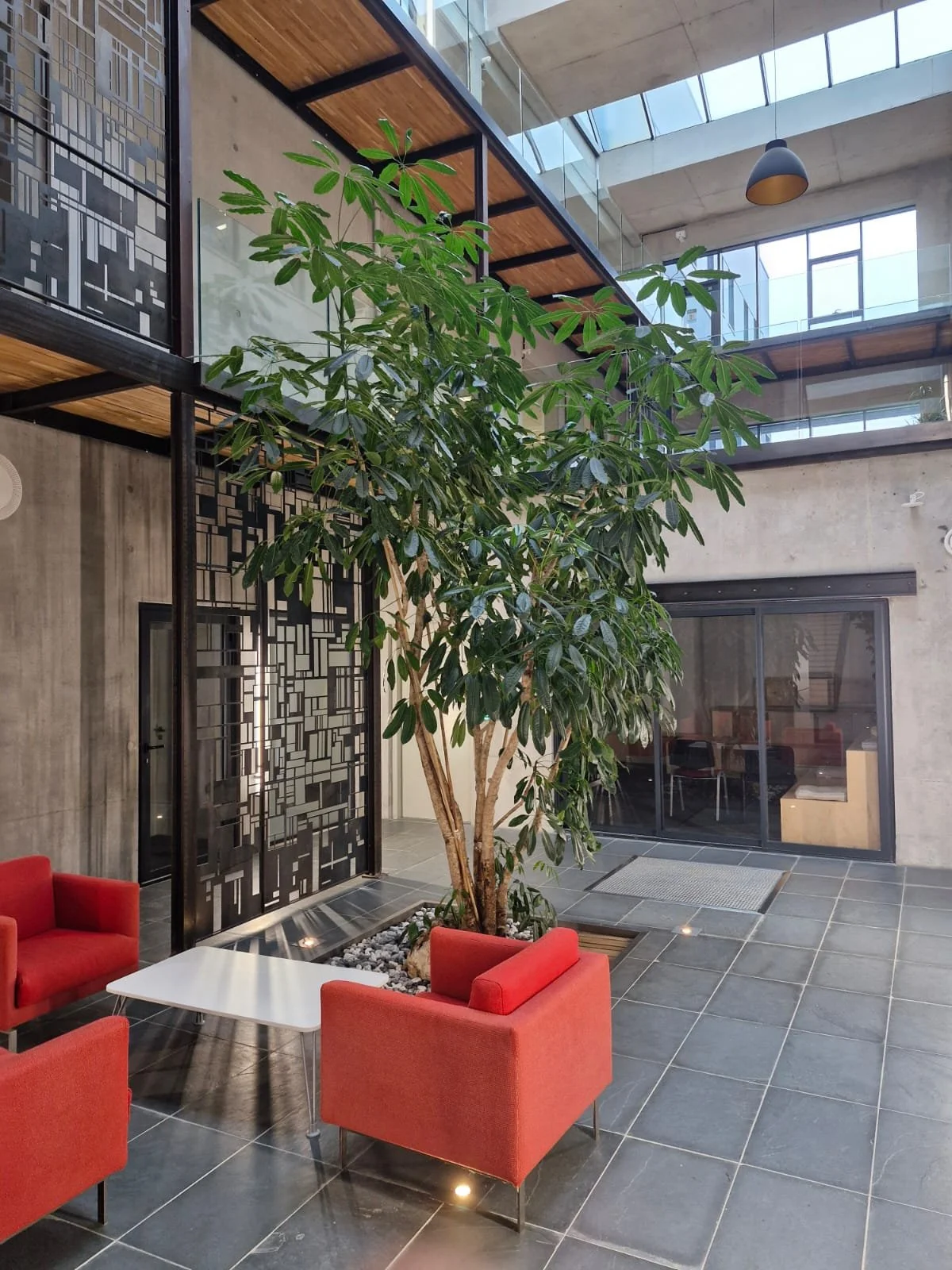 Un hall de bâtiment moderne avec un arbre d'intérieur, fauteuils rouges, une table blanche, murs en béton, fenêtres en verre et un plafond en béton avec des fenêtres en hauteur.