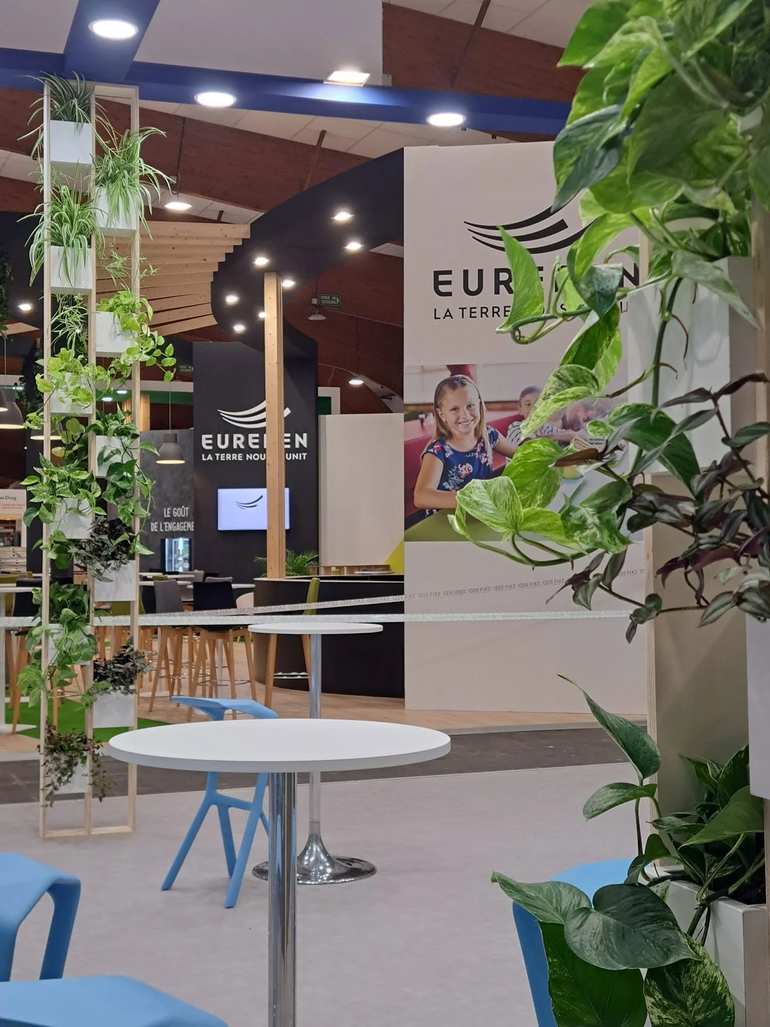 Un espace intérieur moderne avec des plantes vertes en pots et des tables rondes avec des chaises bleues, panneaux d'affichage avec le logo 'EUREN' et une affiche avec une photo d'une fille souriante.