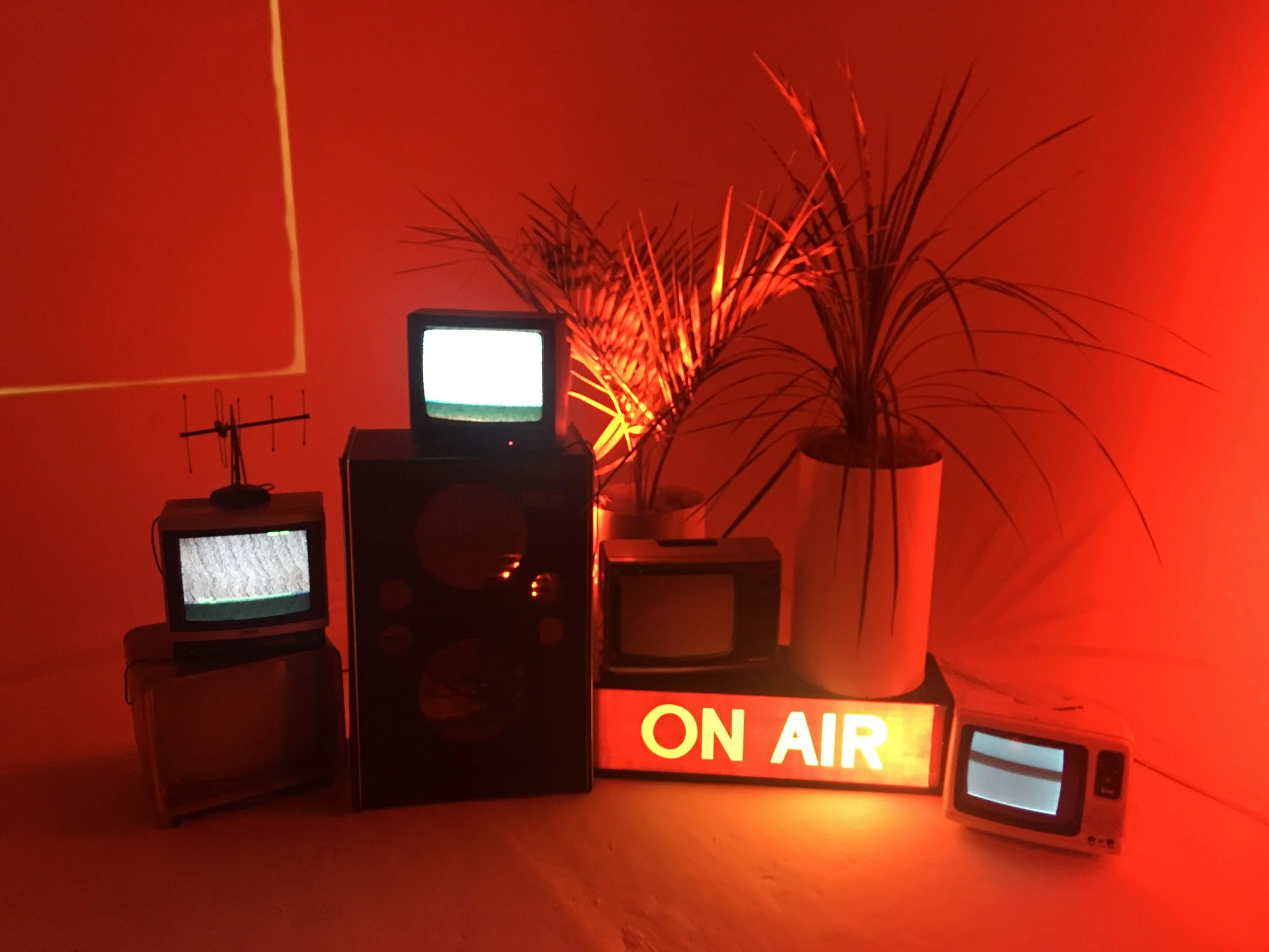 Un espace lumineux rouge avec plusieurs petits téléviseurs anciens affichant un signal brouillé, un panneau lumineux rouge-orange indiquant 'ON AIR', et des plantes en pot.
