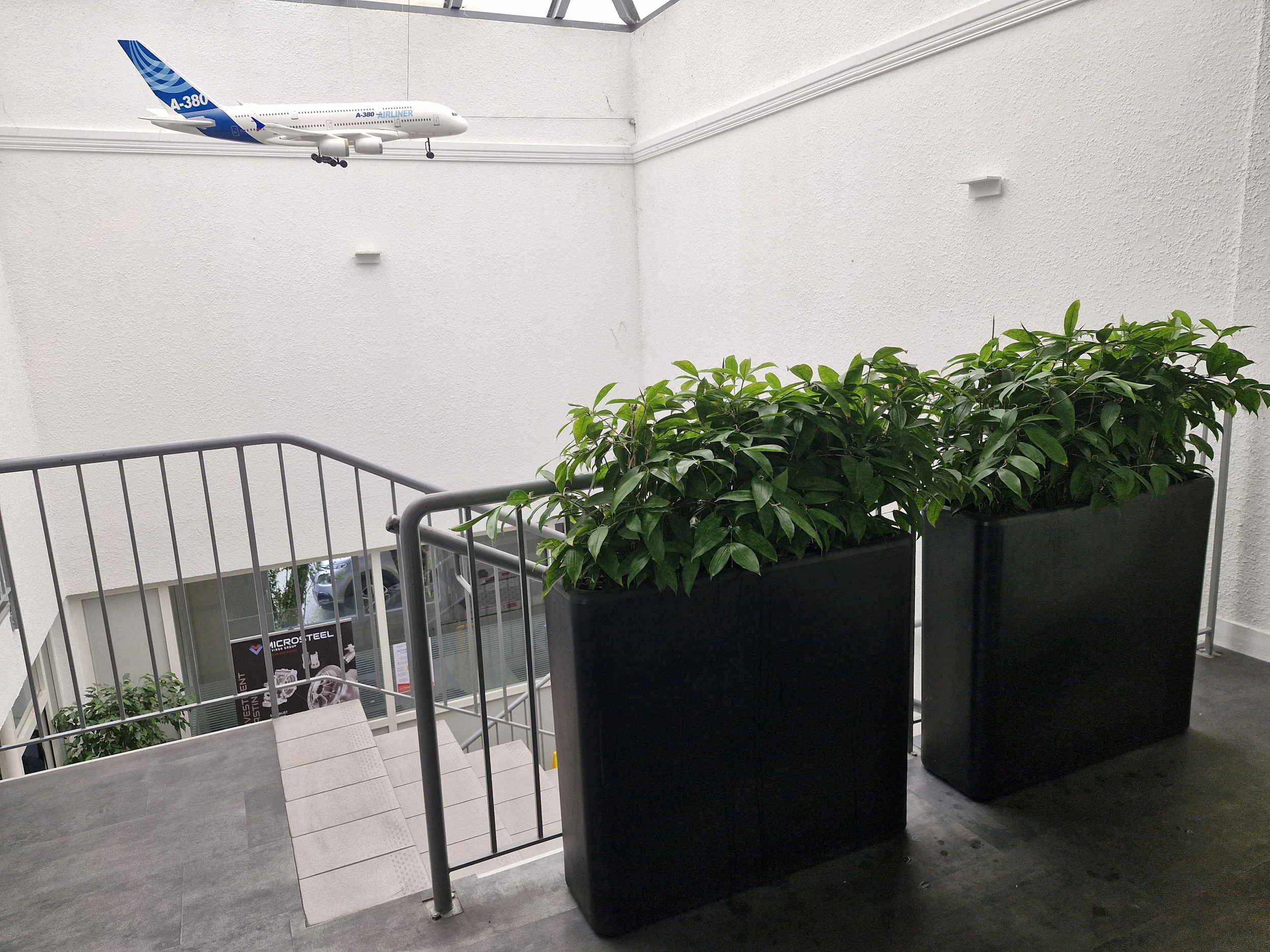 Une reproduction d'un avion Airbus A380 suspendu au plafond d'un bâtiment, avec une grande plante en pot à l'avant, une rampe d'escalier en métal et un mur blanc texturé en arrière-plan.