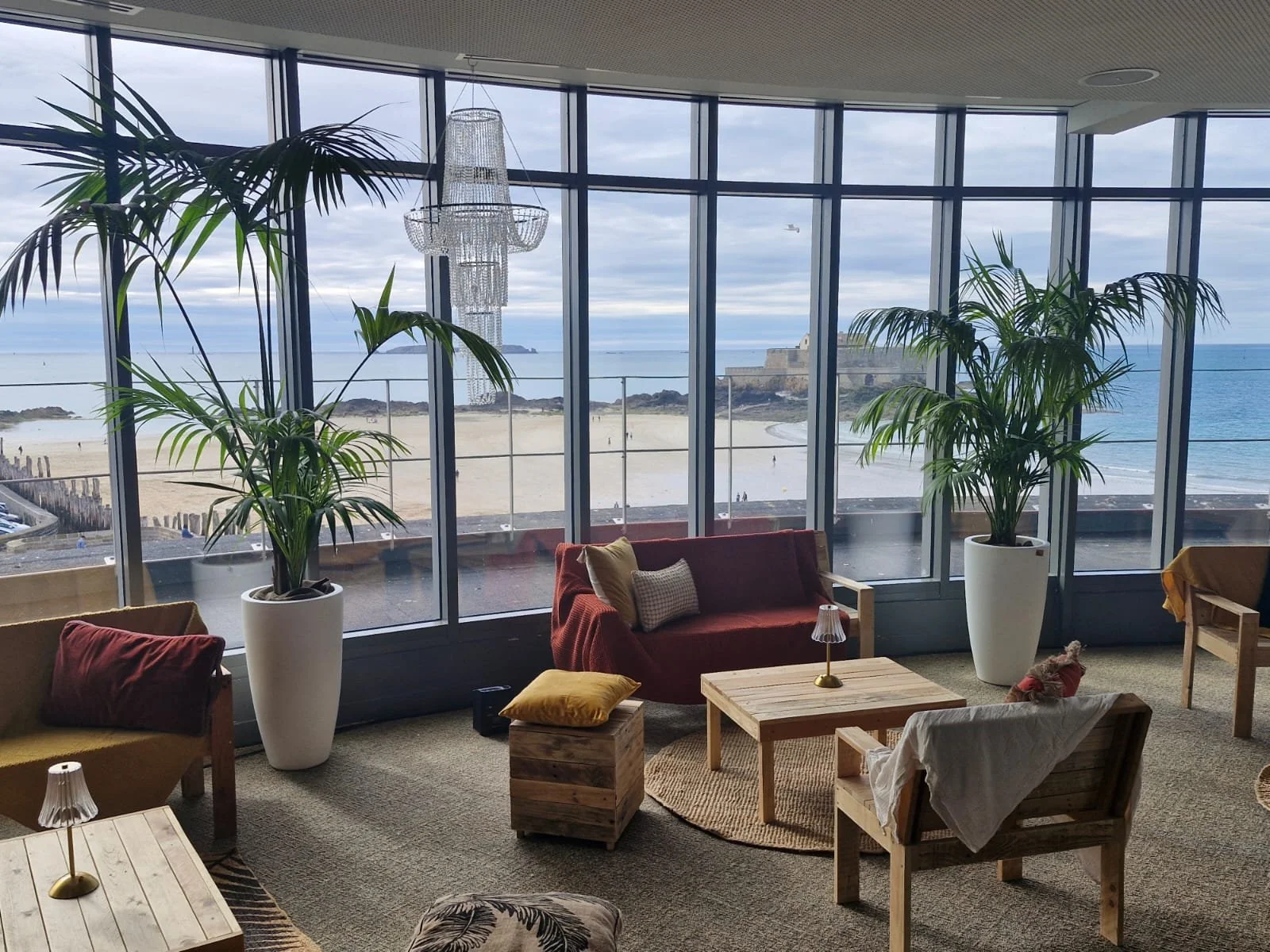 Salon avec grandes fenêtres offrant une vue sur la plage, deux grands meubles avec des plantes en pot, un canapé rouge avec des coussins, une table en bois, un chandelier en cristal suspendu au plafond, ambiance chaleureuse.