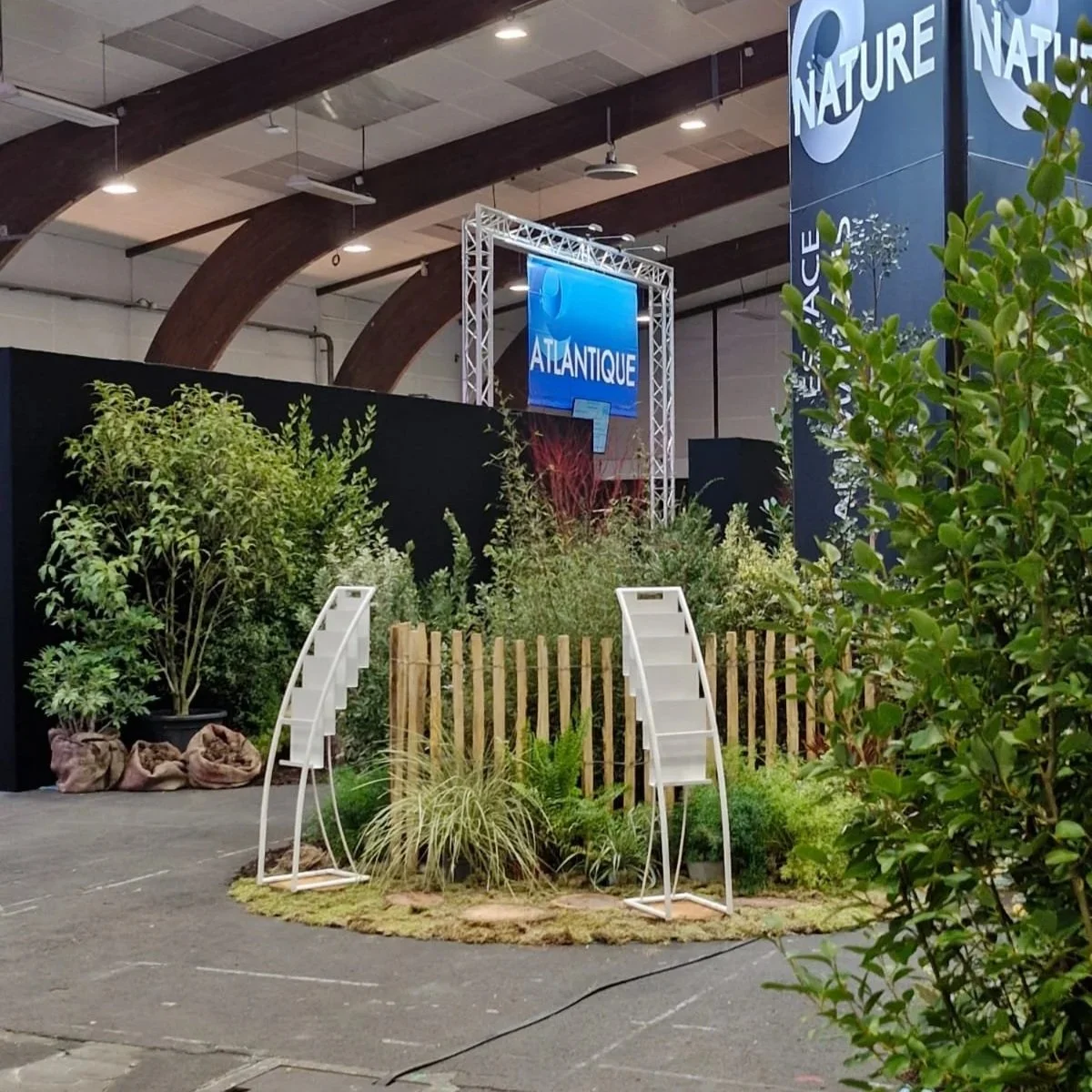 Exposition intérieure avec des plantes, deux petites échelles blanches, une clôture en bois, des panneaux d'affichage bleus avec le mot "Atlantique" et une grande colonne avec le mot "Nature" affiché.