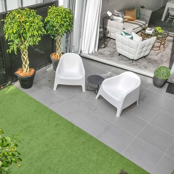 Balcon avec deux fauteuils blancs, une petite table noire, deux grands arbres en pot, un faux gazon vert et un espace intérieur visible à travers une porte-fenêtre avec un salon avec canapé et fauteuils.