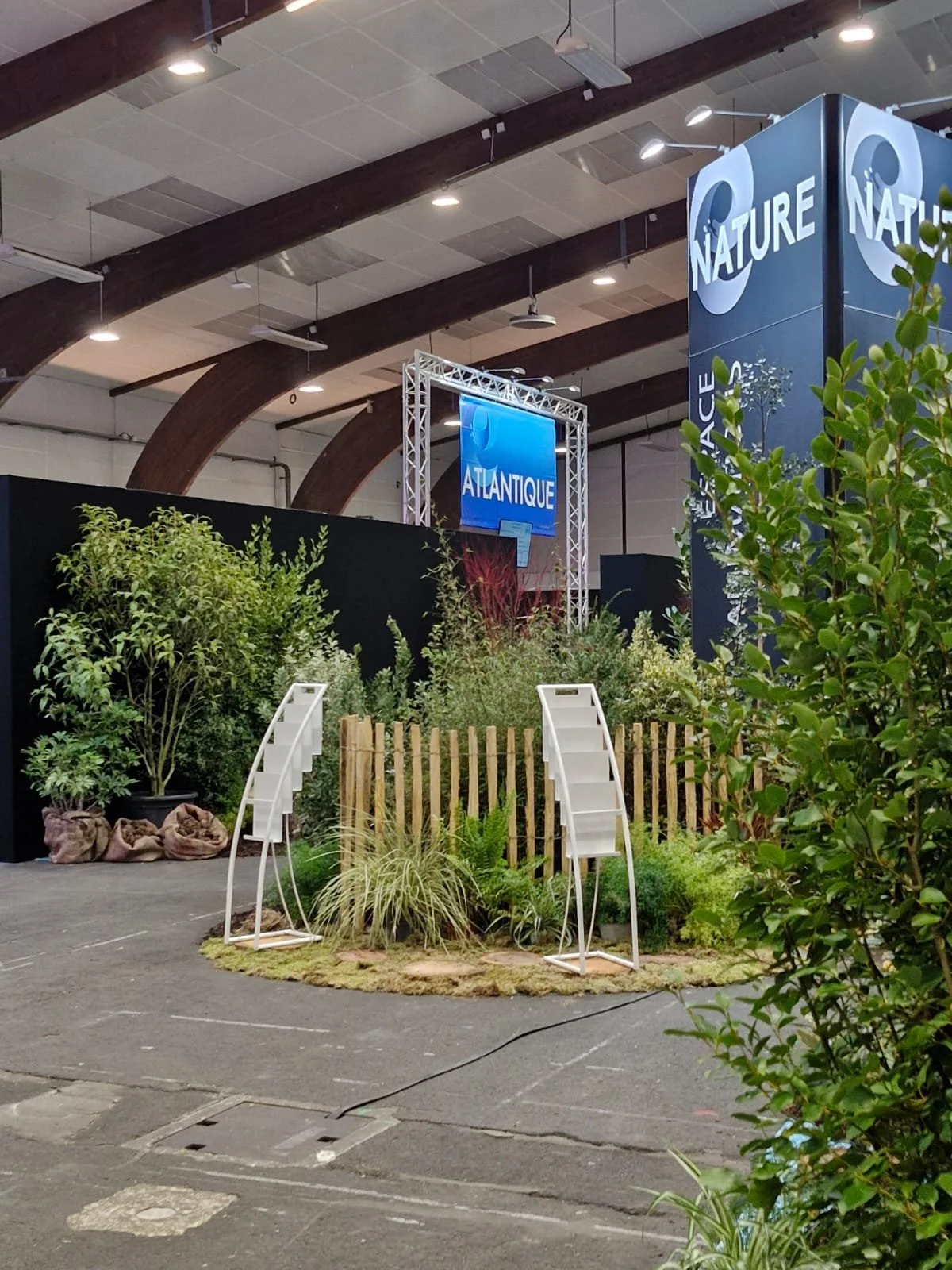 Exposition avec un décor de plantes et de green, deux escaliers blancs, panneaux d'affichage avec les mots "NATURE" et "ATLANTIQUE", dans un intérieur avec poutres en bois et éclairage suspendu.