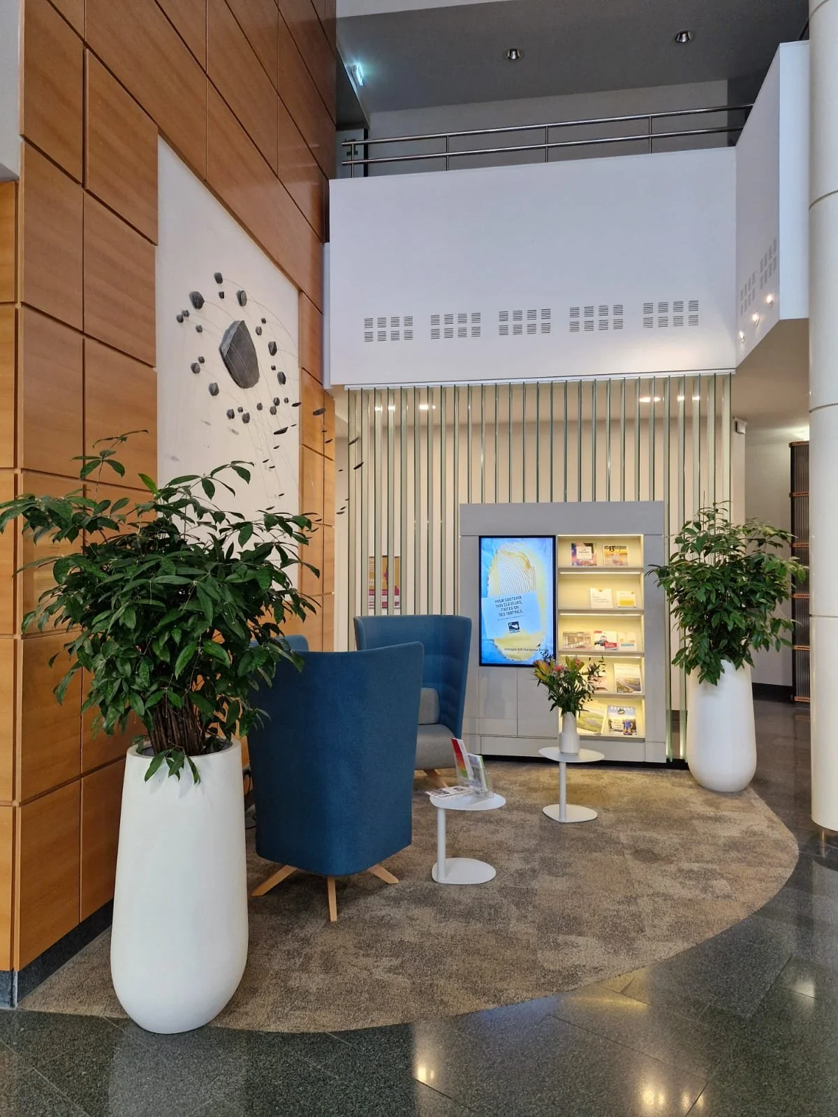 Intérieur d'un espace d'accueil ou de lobby avec deux grands pots blancs contenant des plantes vertes, deux fauteuils bleus, une petite table blanche avec des fleurs, une affiche numérique et une étagère avec des livres, dans un décor moderne avec des murs en bois et en blanc.