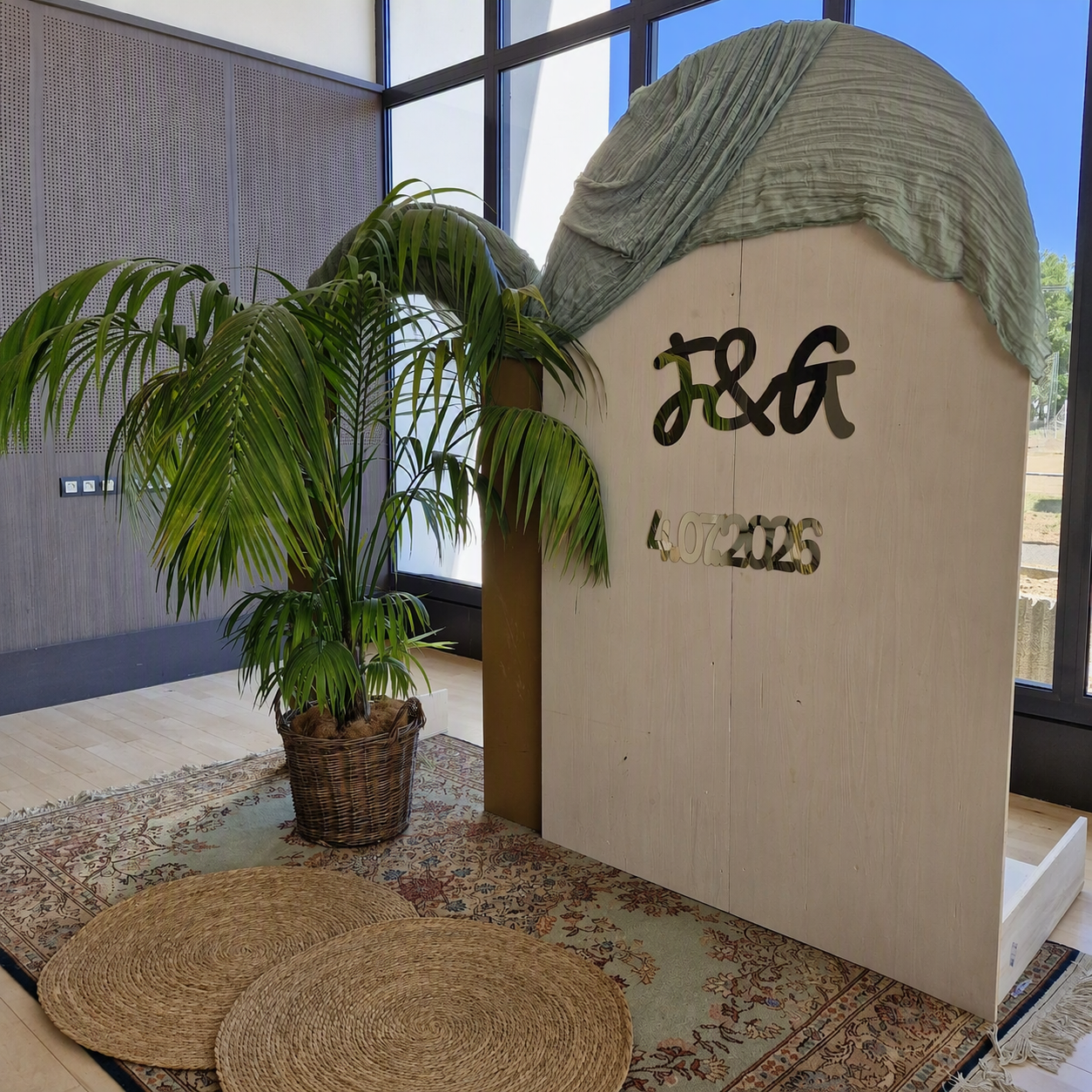 Décor intérieur avec une grande plante en pot, un tapis tissé rond, un panneau en bois clair avec inscription en métal noir et en relief, dans une pièce lumineuse avec de grandes fenêtres.