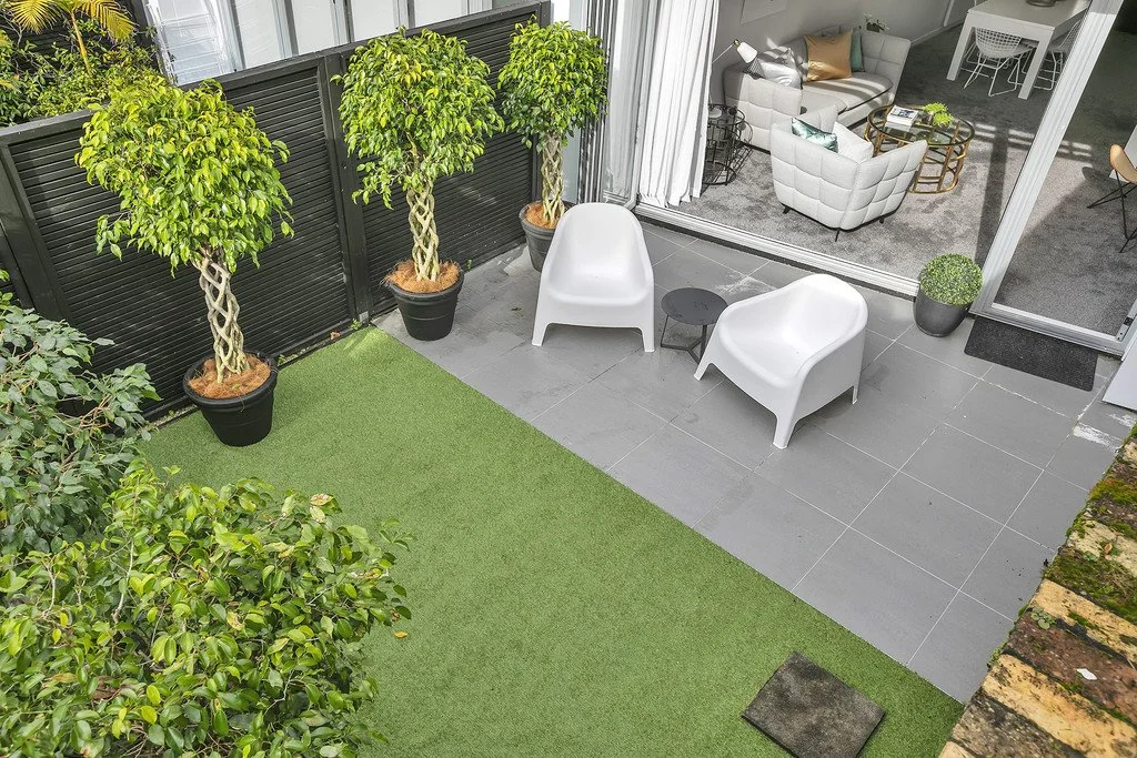 Terrasse avec deux fauteuils blancs, une petite table noire, et des plantes en pots, donnant sur une pelouse artificielle et une pièce intérieure meublée.