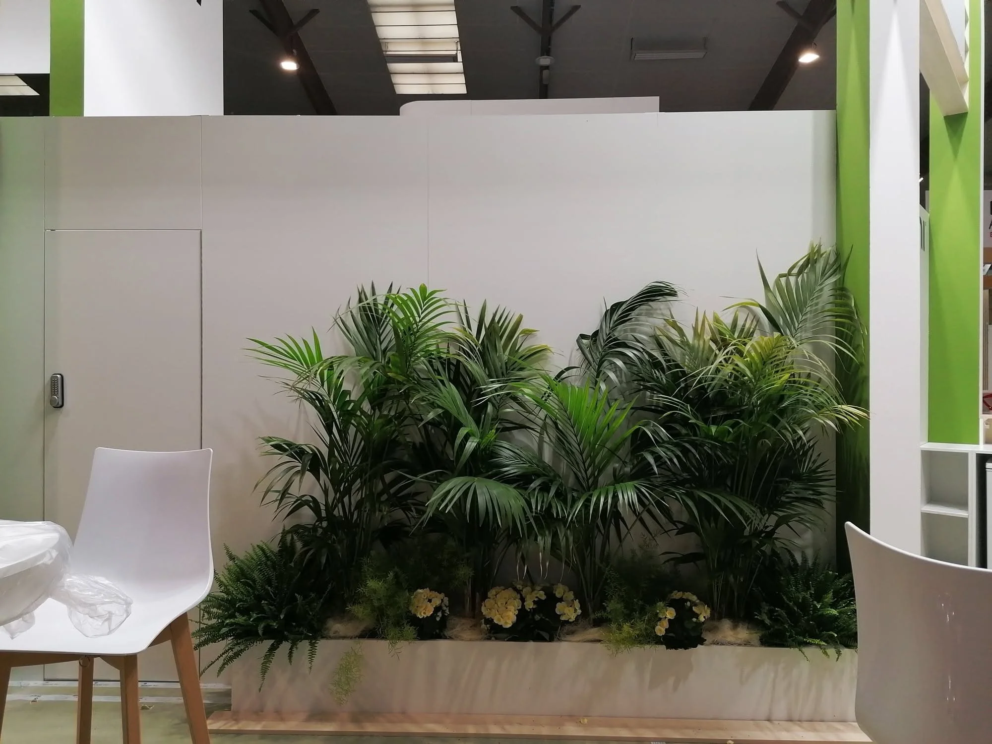 Décoration intérieure avec un arrangement de plantes vertes et des fleurs blanches situées en bas. Mur blanc derrière avec un panneau绿色 à droite et une chaise blanche à gauche.
