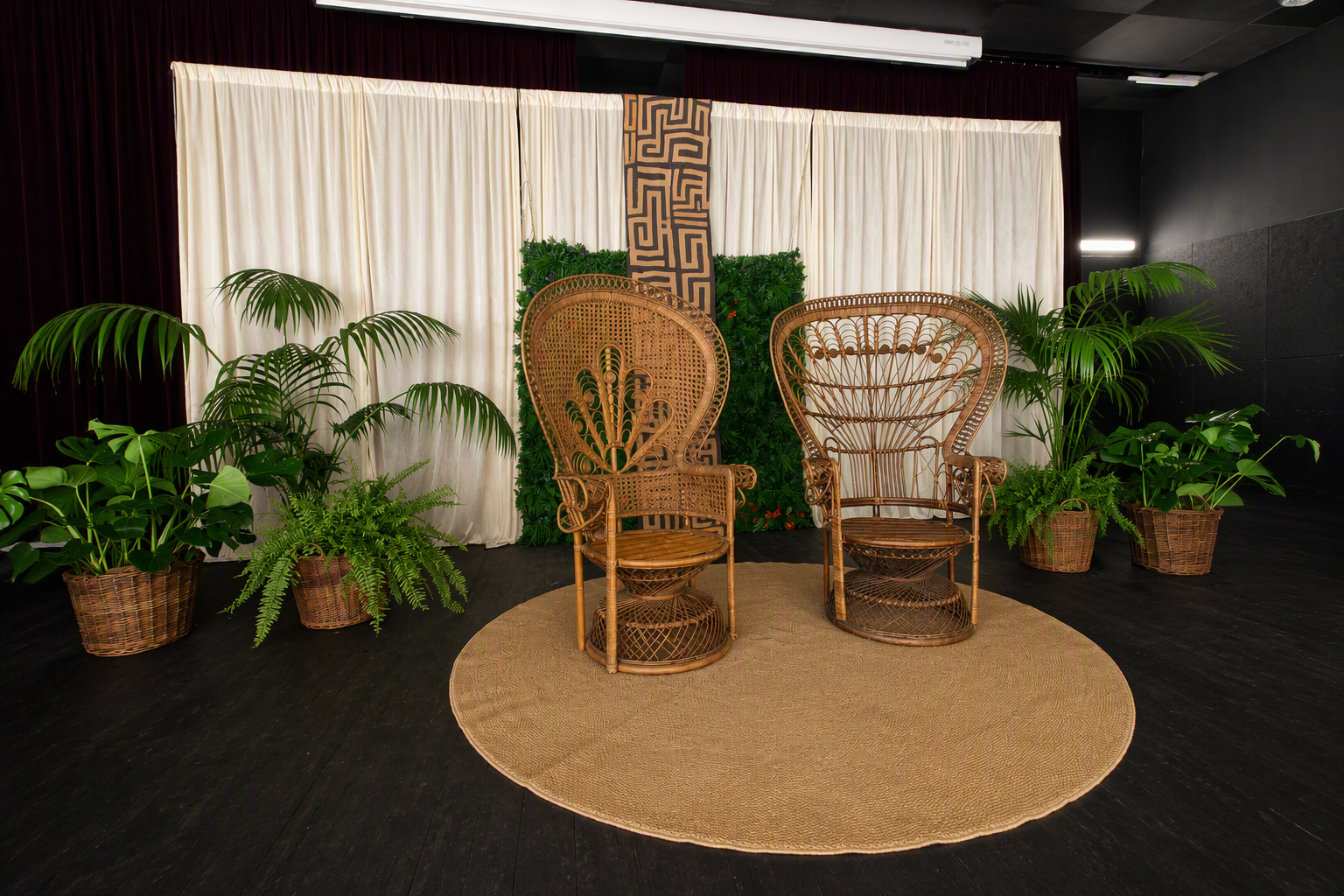 Deux chaises en rotin anciennes face à face sur un tapis rond en bambou, entourées de plusieurs plantes vertes en pot et d'un fond de rideaux blancs avec un panneau décoratif.
