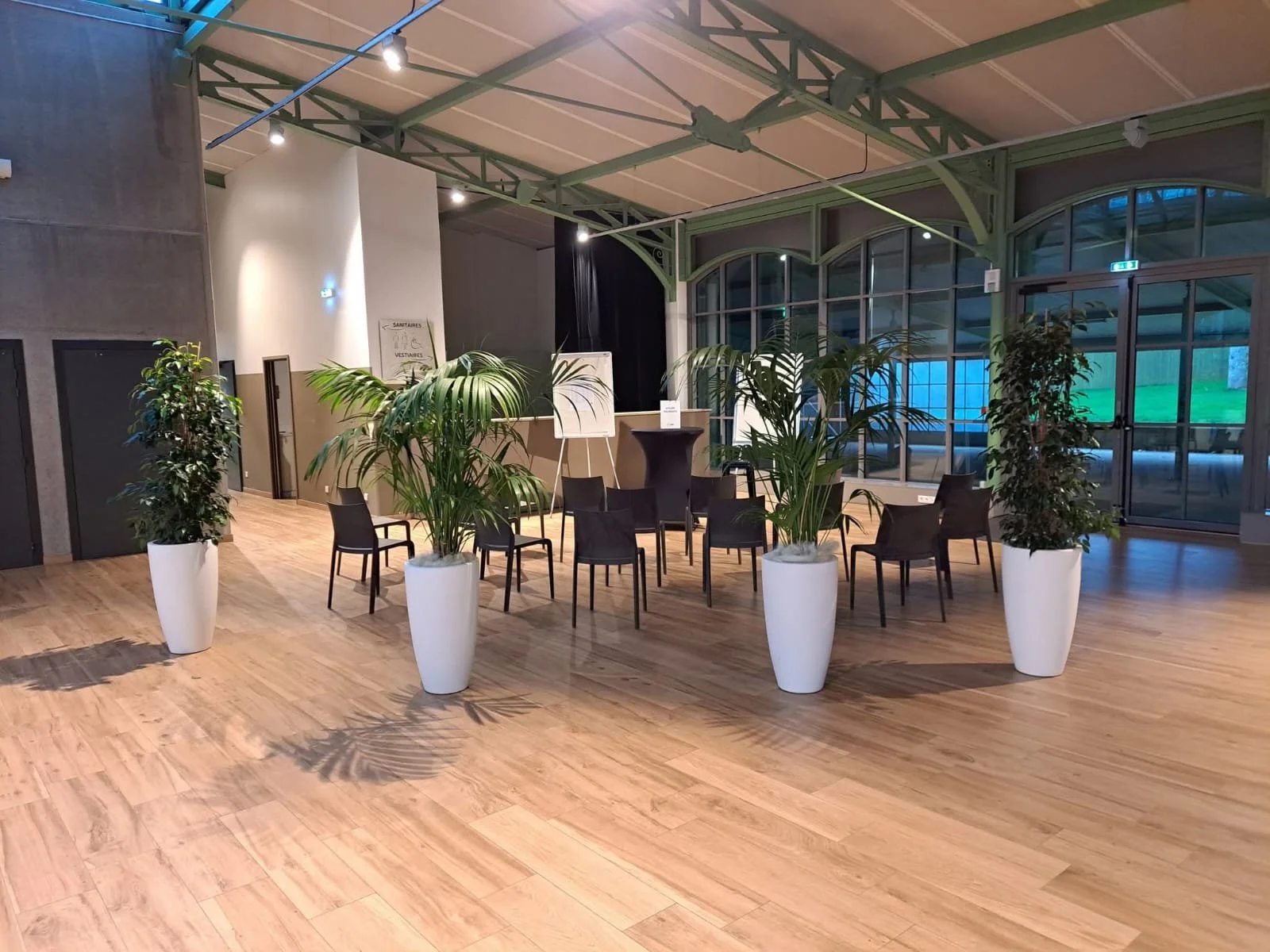Salle d'événement intérieure avec des chaises noires disposées en cercle, entourée de grandes plantes vertes en pots blancs, avec un sol en bois clair et des fenêtres en verre laissant entrer la lumière naturelle.