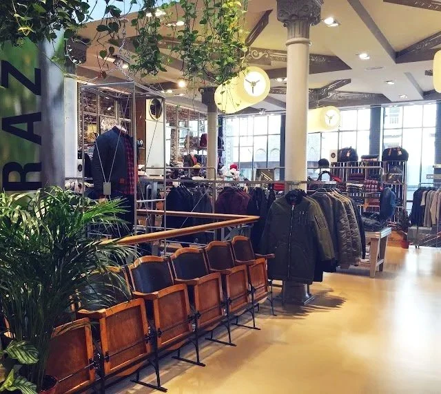 Intérieur d'un magasin de vêtements avec des chandails et manteaux suspendus, des rangées de chaises en bois, des plantes vertes, et de grandes fenêtres permettant à la lumière naturelle de pénétrer.