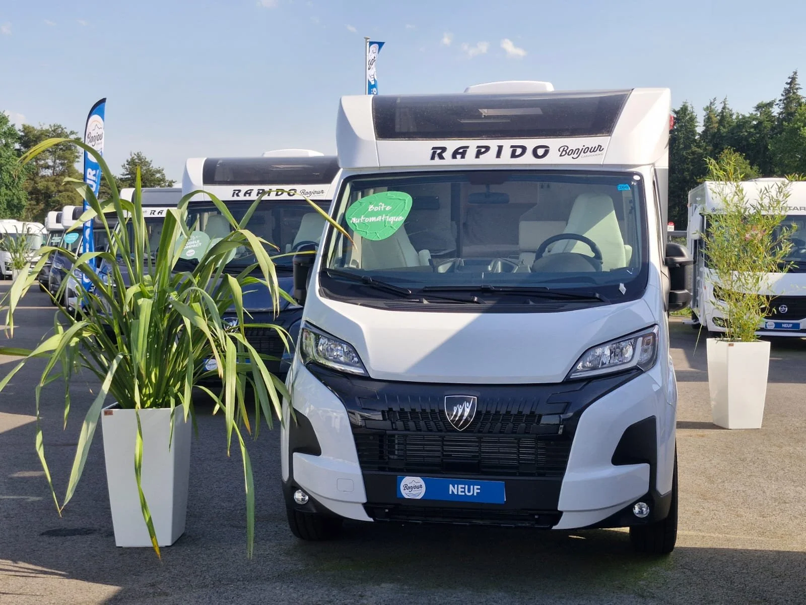 Voiture neuve blanche de marque Rapid'O Bonjour, exposée en extérieur, avec deux grands pots de plantes vertes de chaque côté, sous un ciel clair.