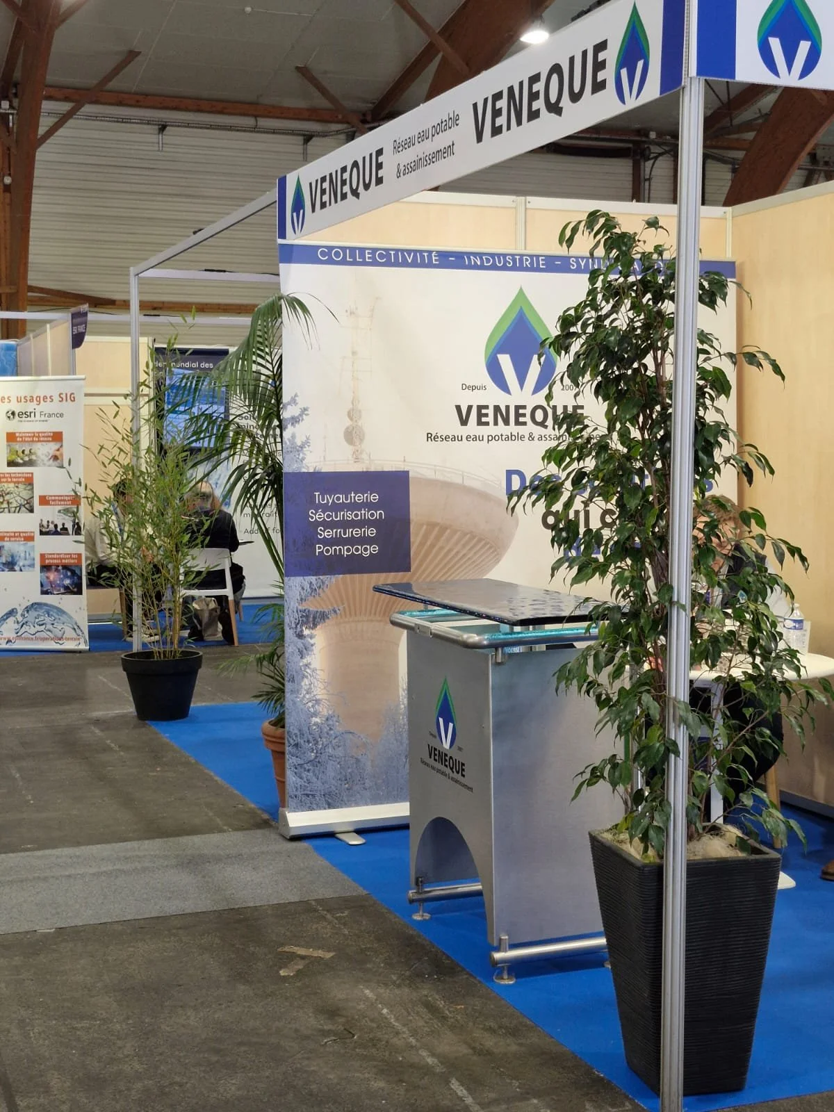 Stand d'exposition de la société Venque, spécialisé dans les réseaux d'eau potable et d'assainissement, avec des plantes en pot et une bannière illustrant leurs services.