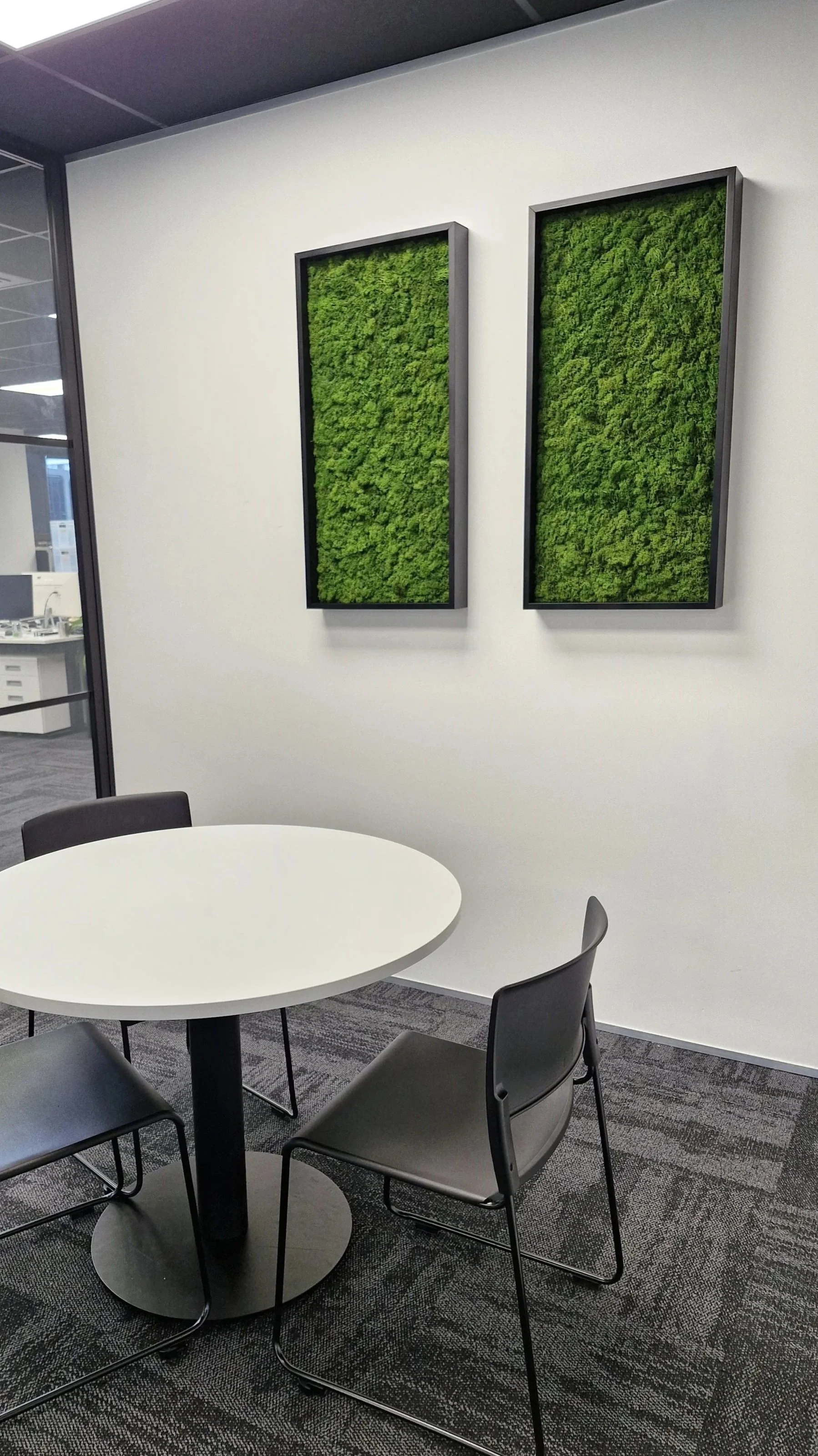 Salle de réunion moderne avec un tableau blanc, une table ronde blanche entourée de quatre chaises en plastique noir, et deux cadres avec de la verdure suspendus au mur blanc, dans un environnement de bureau.