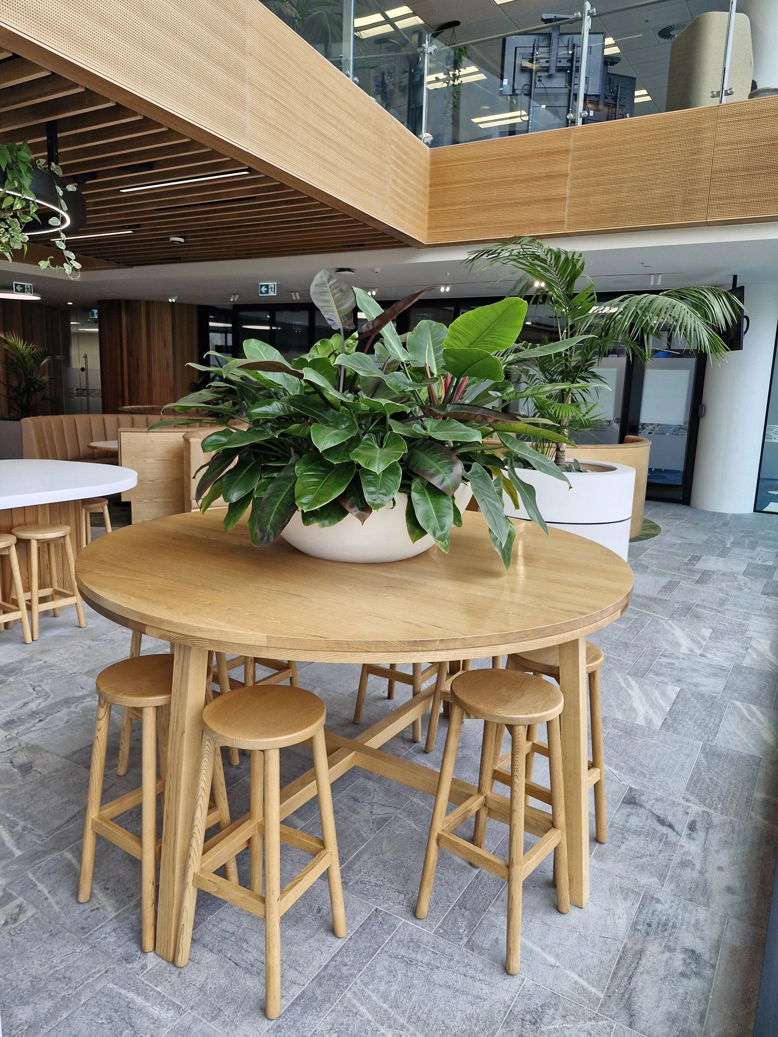 Une table en bois avec des tabourets en bois autour, décoration intérieure moderne avec plantes vertes et mobilier en bois clair, dans un espace lumineux avec une mezzanine et des fenêtres en arrière-plan.