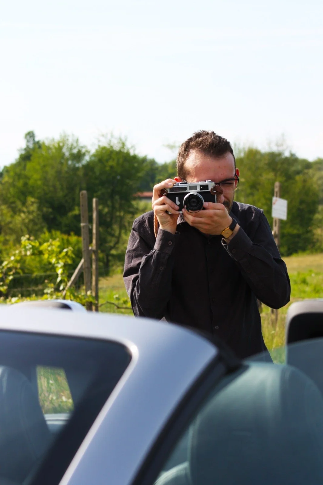 Uomo che scatta una foto con una fotocamera digitale in un'area aperta, con auto in primo piano e vegetazione sullo sfondo.