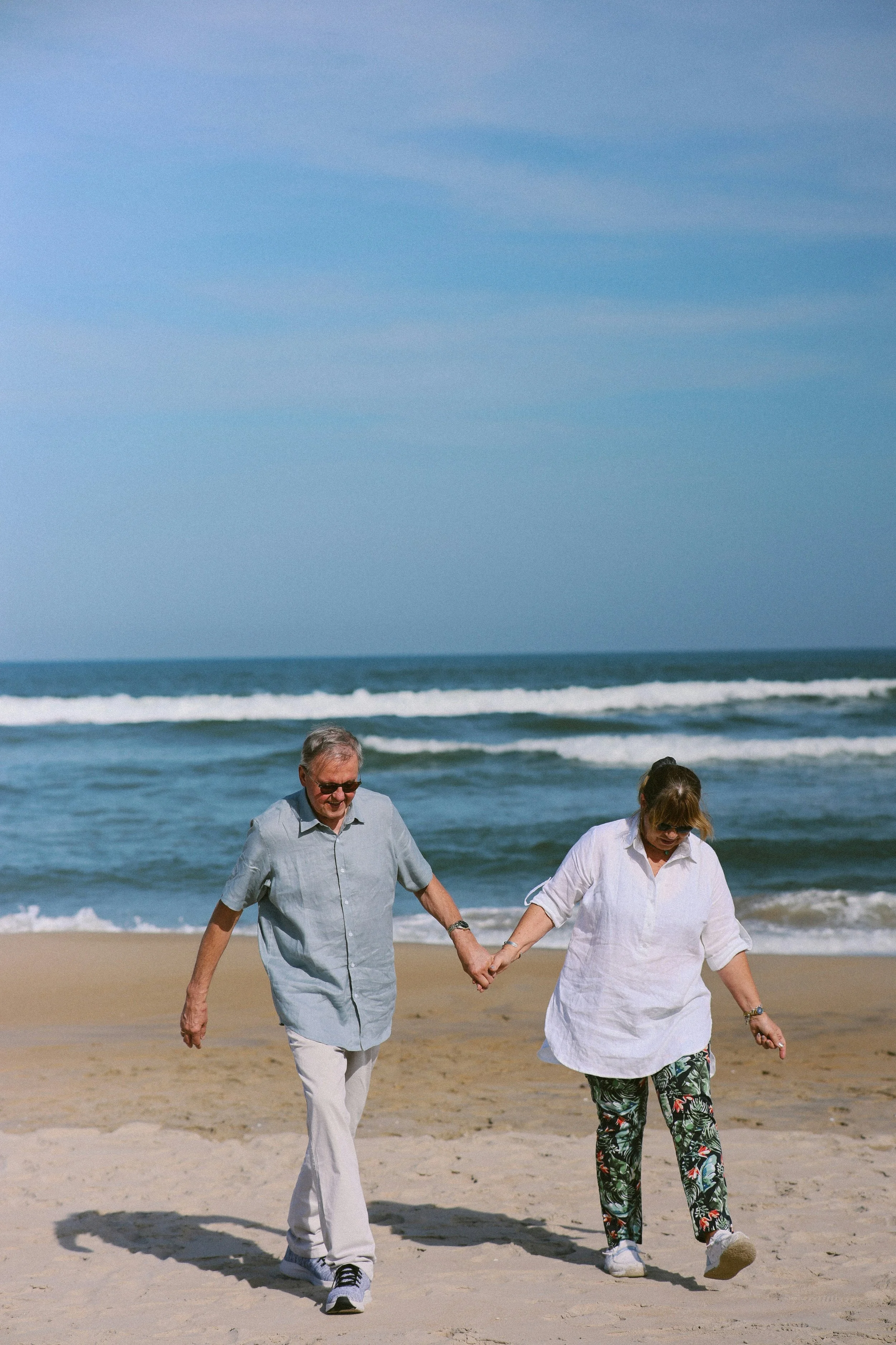 A man and woman walking hand in hand on the beach, close to the shoreline, with the ocean waves and blue sky in the background.