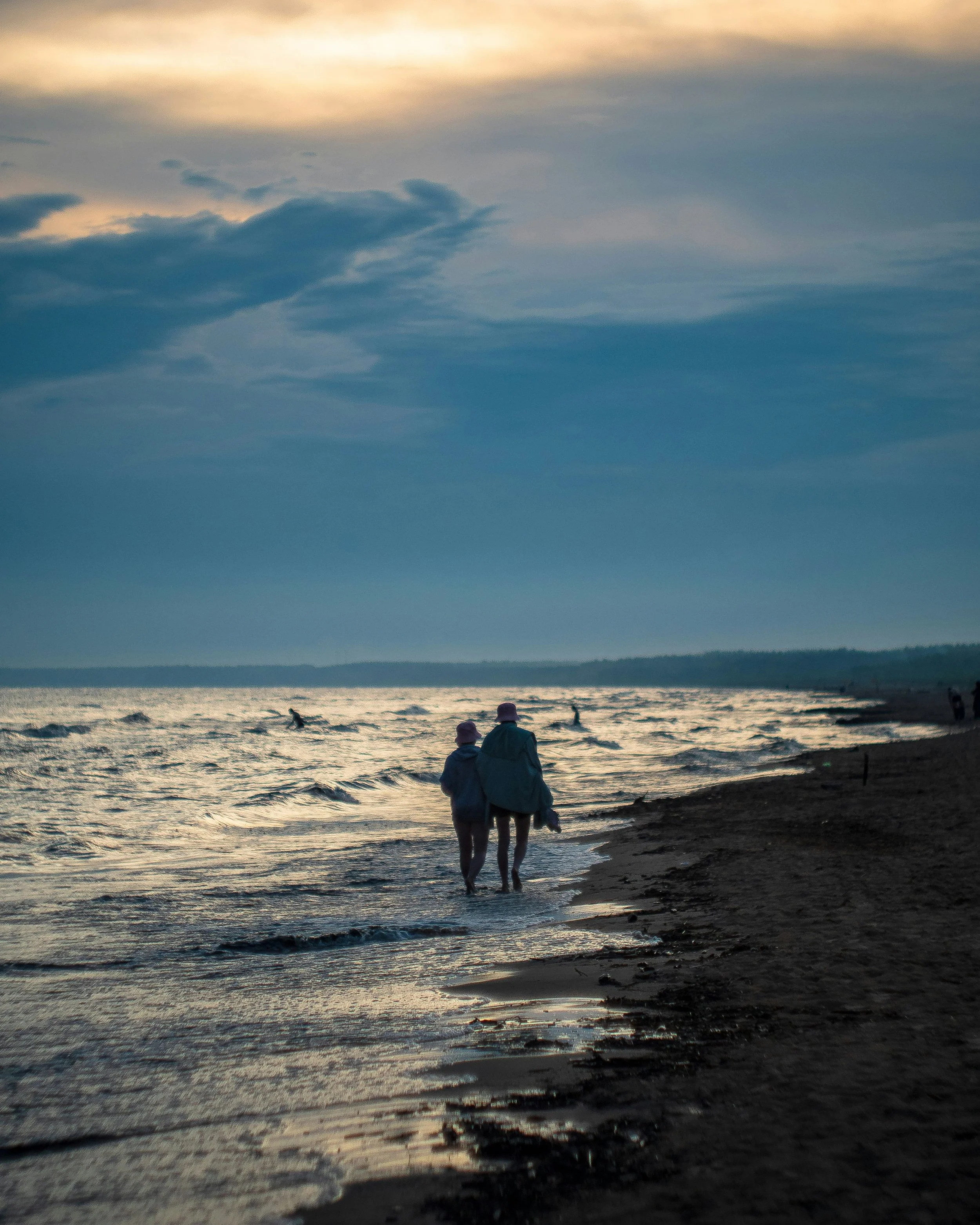 People walking along the shoreline at sunset or dusk, with others surfing in the water, and cloudy sky overhead.
