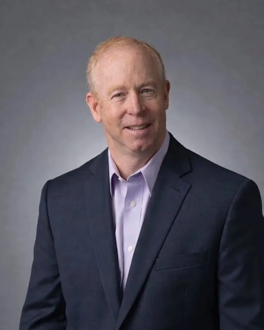 A man in a dark suit and light purple shirt smiling against a gray background.