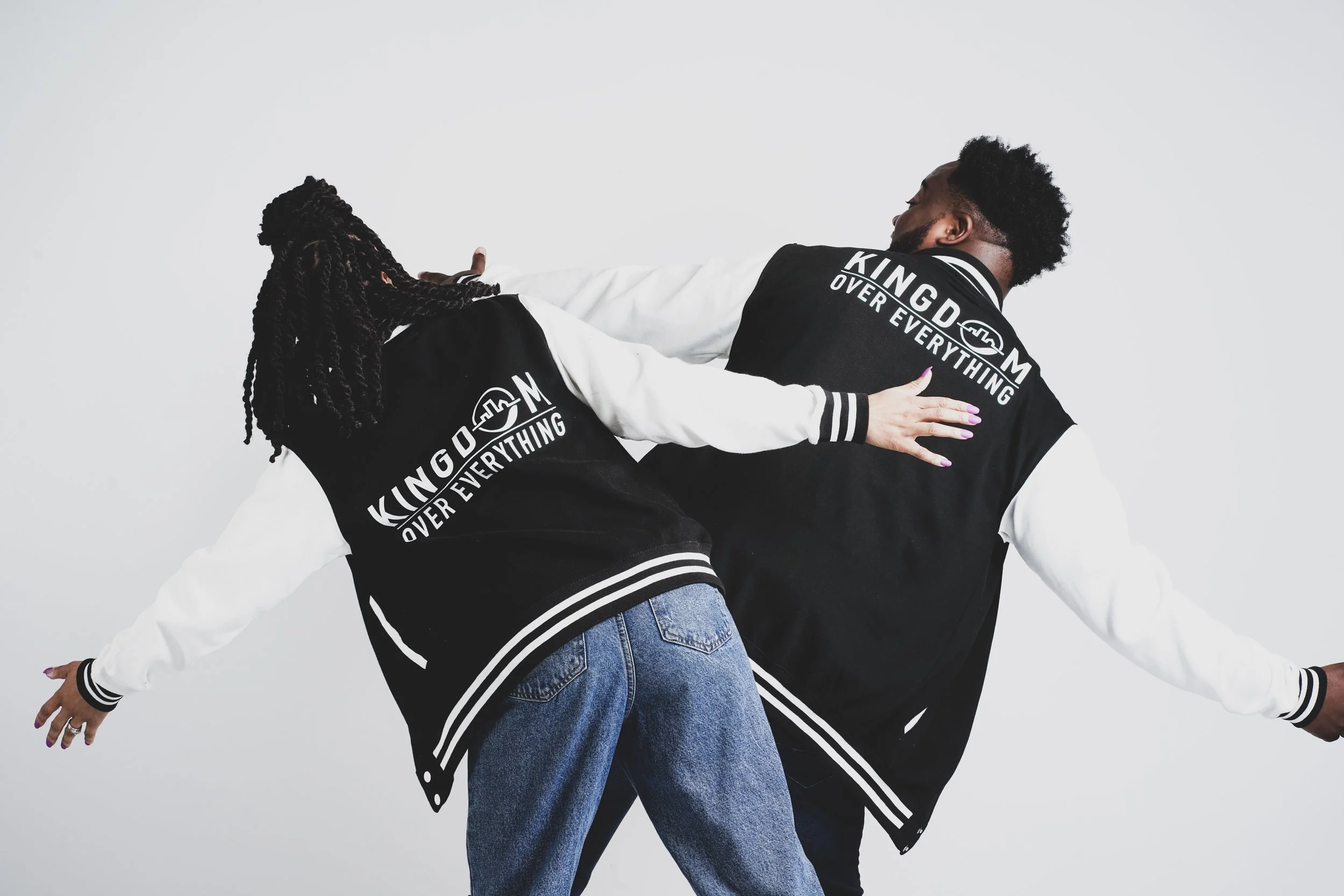 Two people wearing matching black and white varsity jackets with the text 'KINGDOM OVER EVERYTHING' on the back, posing against a plain white background.