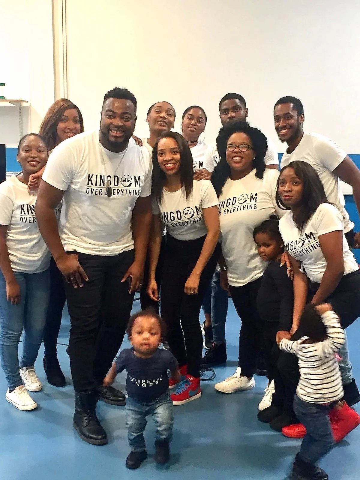 A group of diverse people, including children, posing together in a gym or sports facility. Many are wearing matching white t-shirts with the words 'KINGDOM OVER EVERYTHING'. They are smiling and standing close to each other.