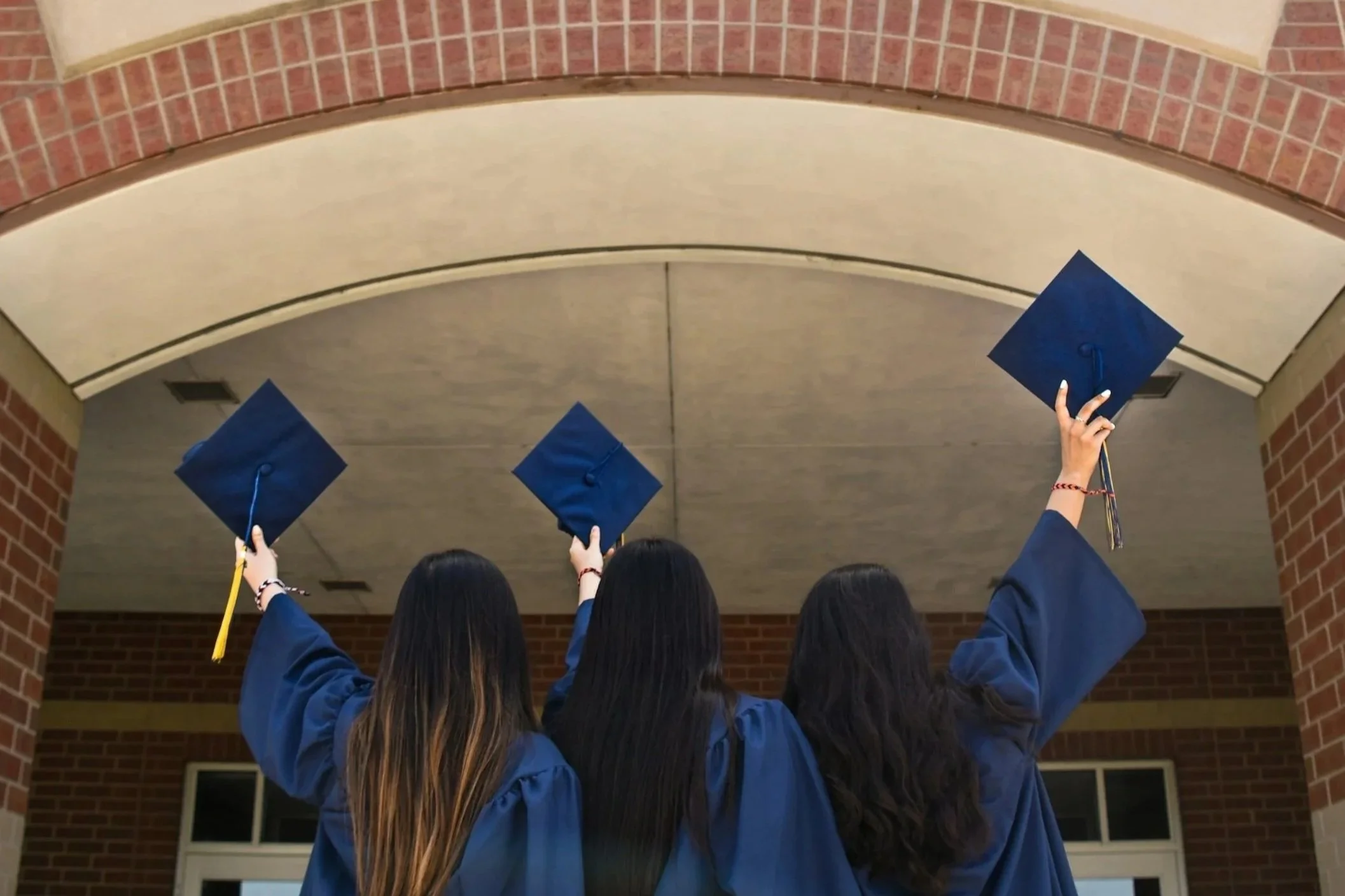 A group of graduates in blue caps and gowns celebrate outdoors, seen from behind, raising their diplomas in the air during a graduation ceremony under an arched brick structure.