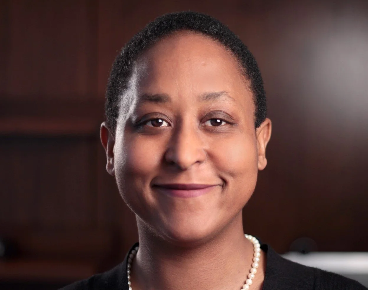 Close-up of an African American woman with short hair smiling, wearing a pearl necklace and black top, against a dark wooden background.