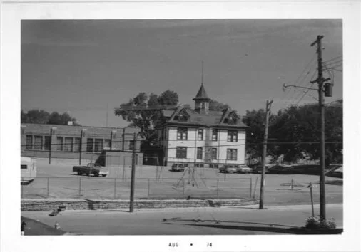 Black and white photo of a vintage-style building with a pointed tower, surrounded by a fence, utility poles, and cars parked nearby, with trees in the background.