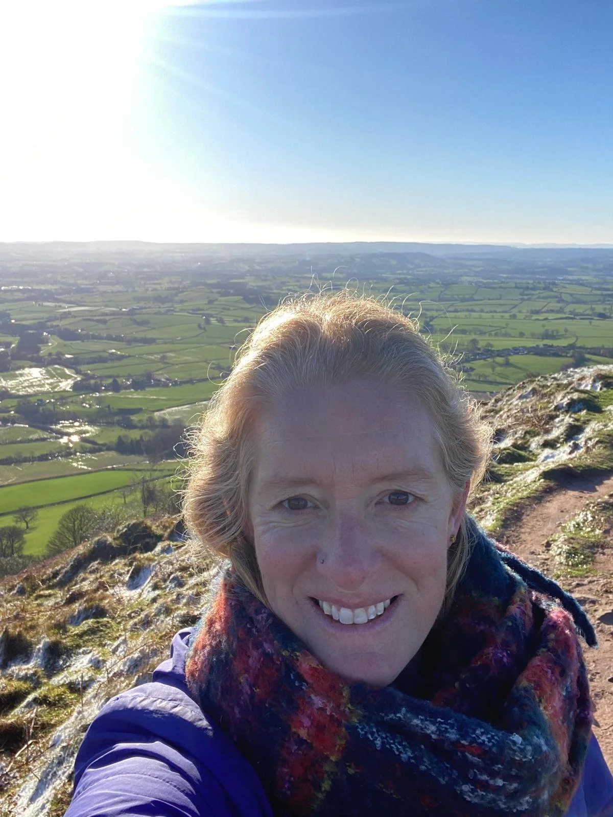 A woman taking a selfie on a mountain trail with a view of green fields and a bright blue sky in the background.