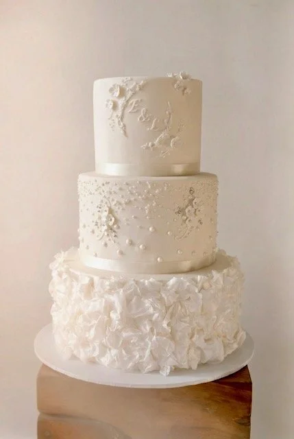 Three-tier white wedding cake with elegant textured decorations, placed on a white plate on a wooden surface.
